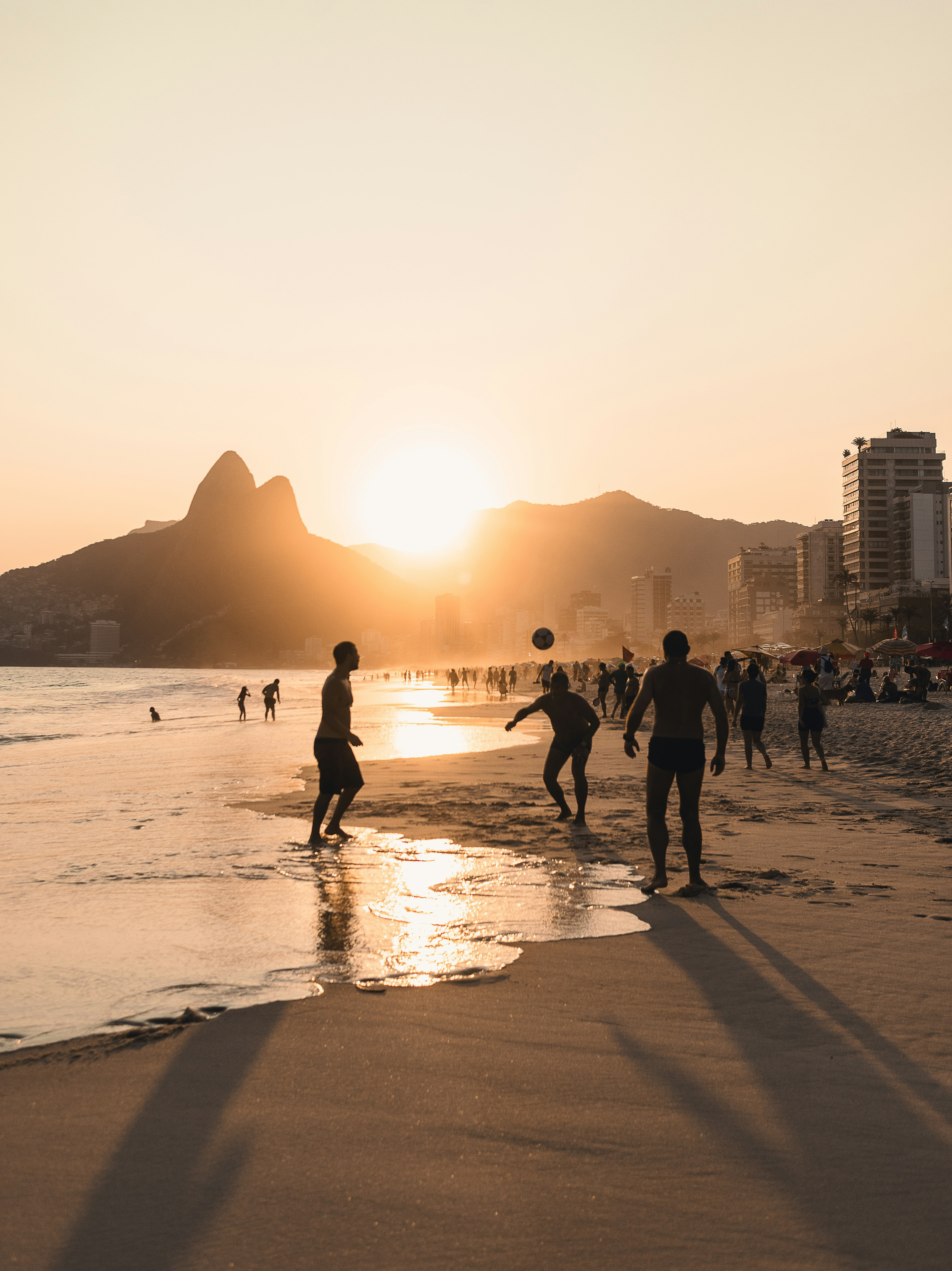 People walk along wet sand at sunset, with orange light reflecting on the beach and a hazy horizon ahead.