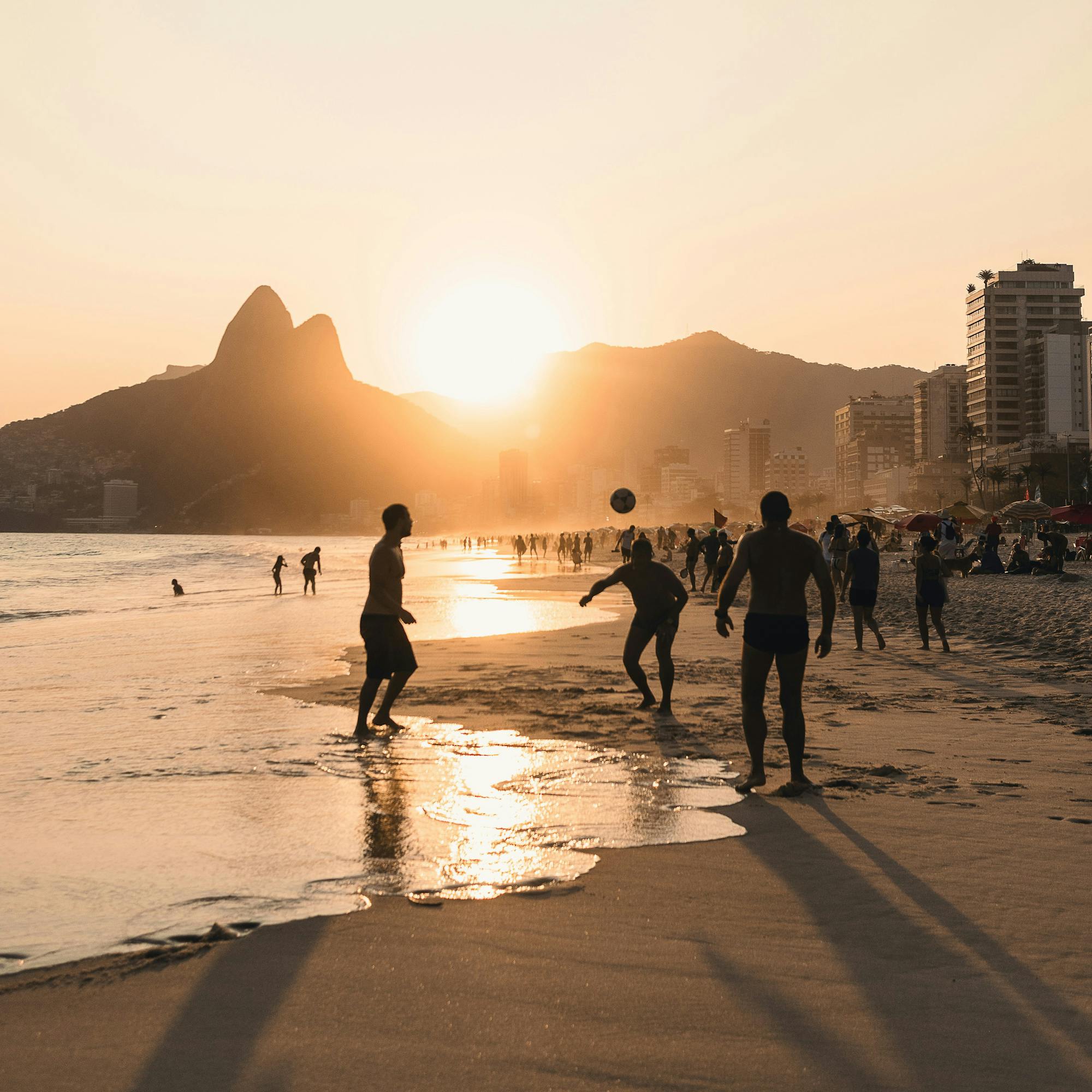 People walk along wet sand at sunset, with orange light reflecting on the beach and a hazy horizon ahead.