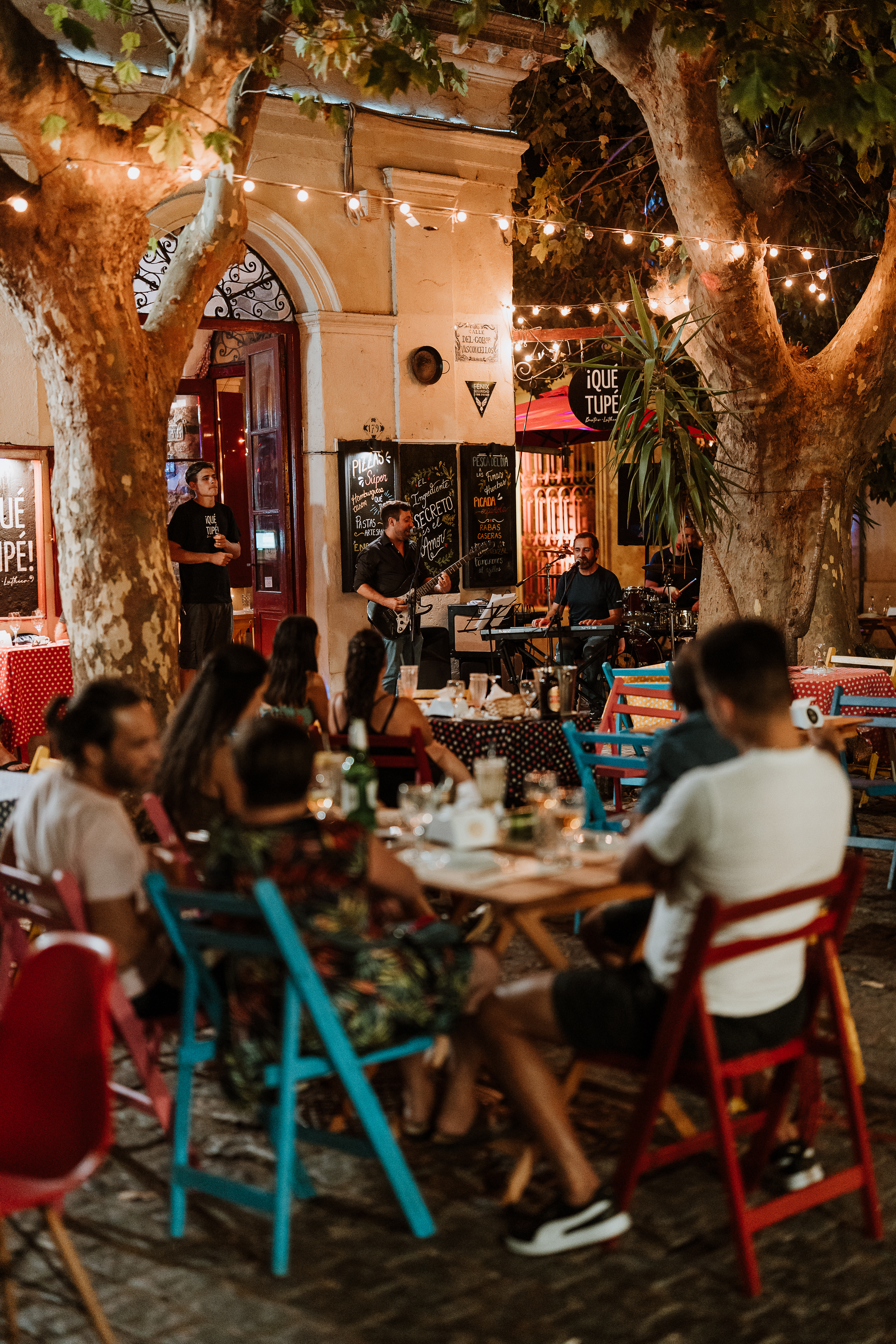 Warm restaurant interior with diners at small tables, surrounded by eclectic decor, hanging lights, and patterned textiles on walls.