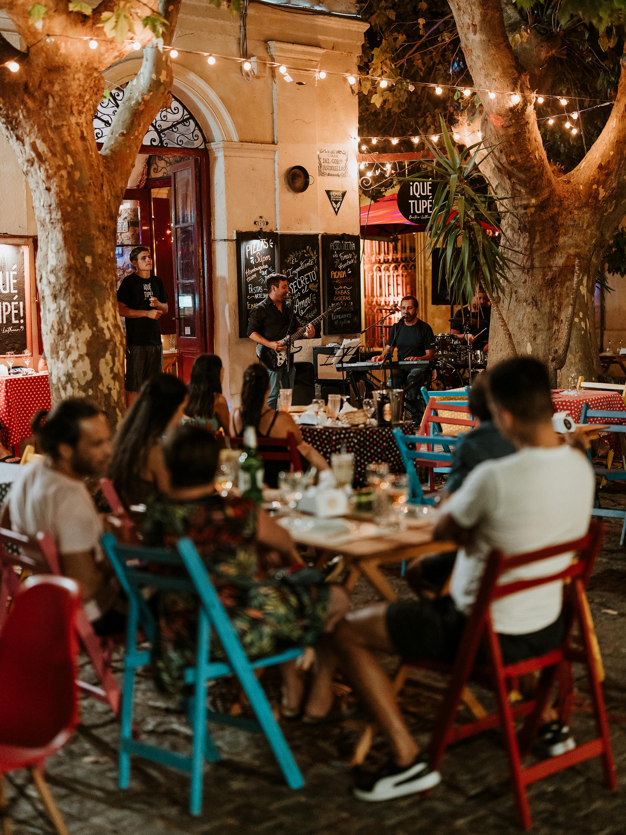 Warm restaurant interior with diners at small tables, surrounded by eclectic decor, hanging lights, and patterned textiles on walls.