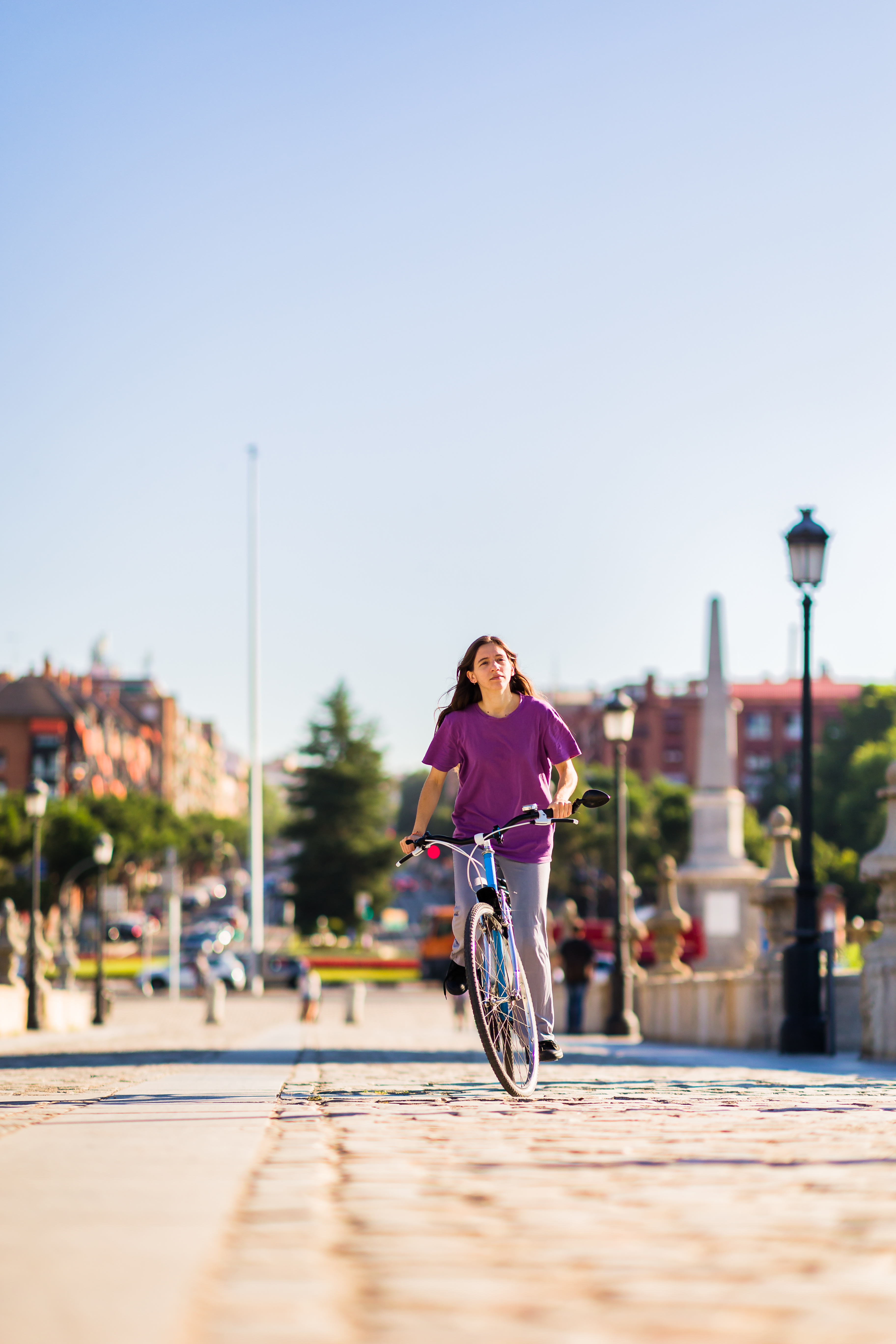 A woman rides a bicycle along a sunlit promenade, with blurred buildings, trees, and lampposts behind her.