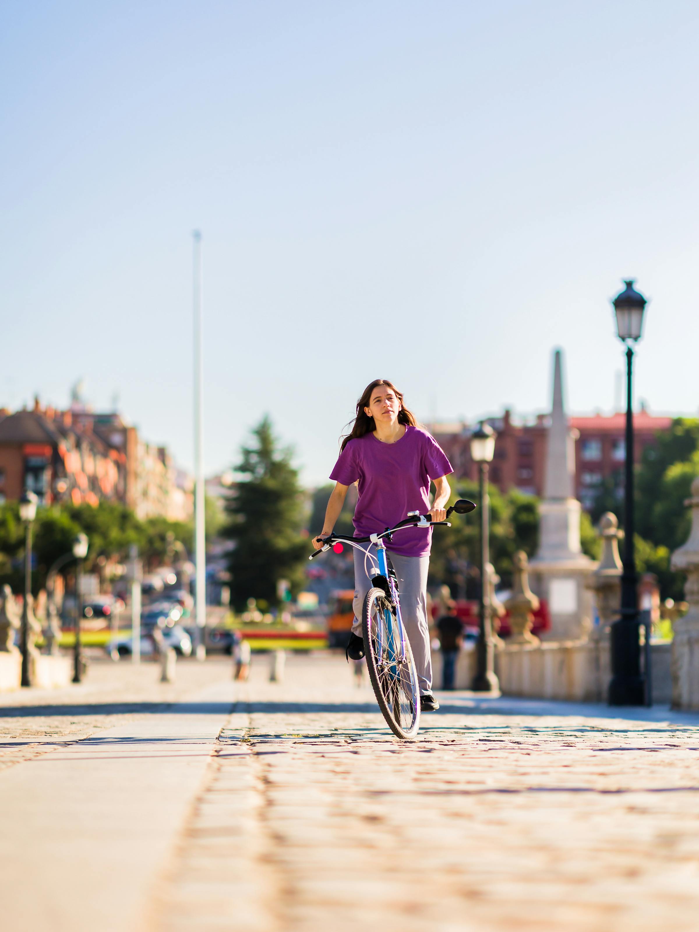 A woman rides a bicycle along a sunlit promenade, with blurred buildings, trees, and lampposts behind her.
