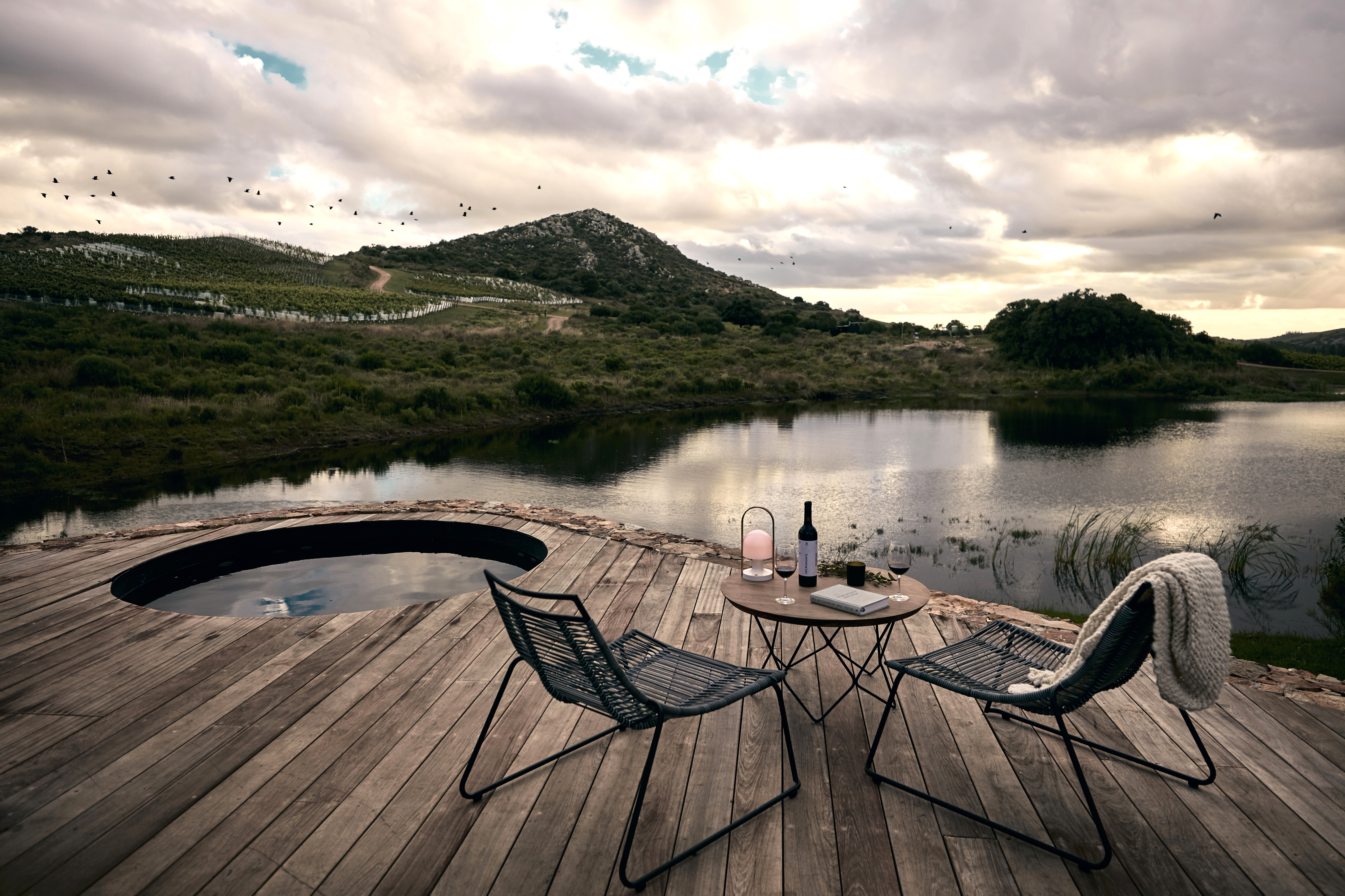 Two lounge chairs rest on a wooden deck beside calm water, with grassy hills and dramatic clouds beyond them quietly.