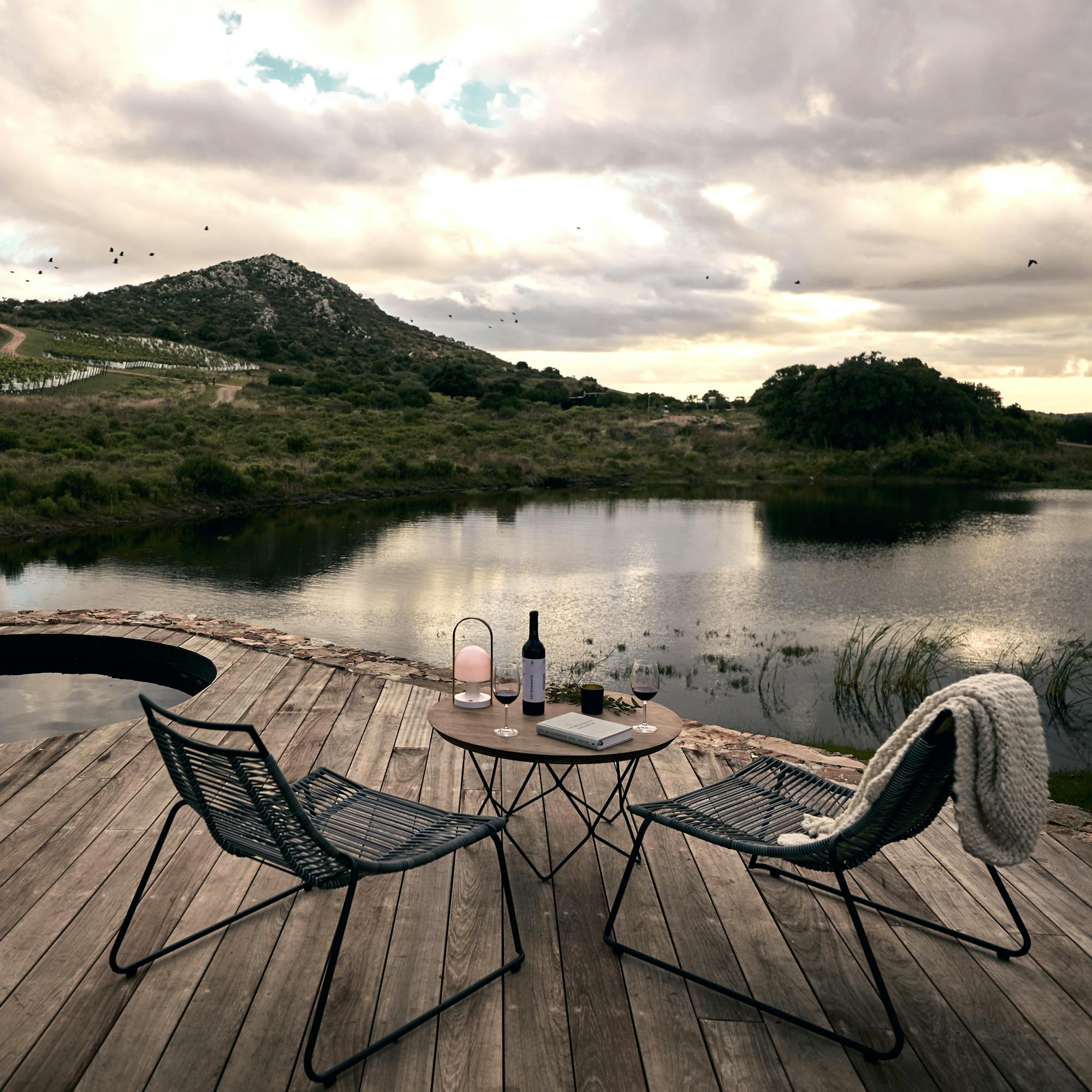 Two lounge chairs rest on a wooden deck beside calm water, with grassy hills and dramatic clouds beyond them quietly.