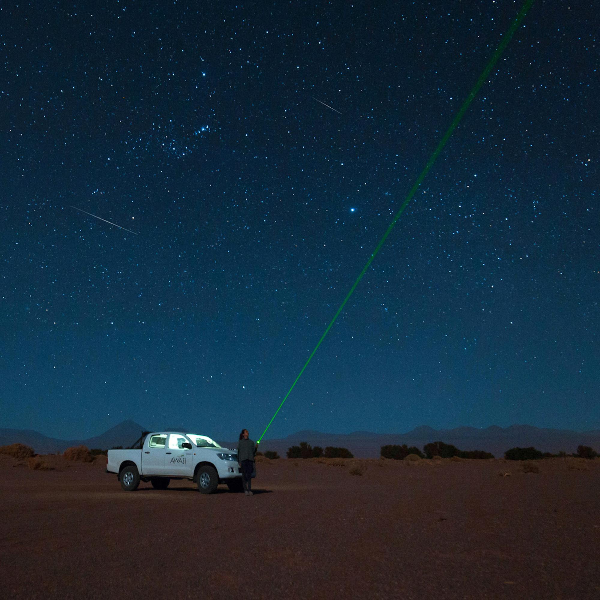Car sits on dark desert sand under a star-filled sky, with a band of Milky Way arching overhead at night.