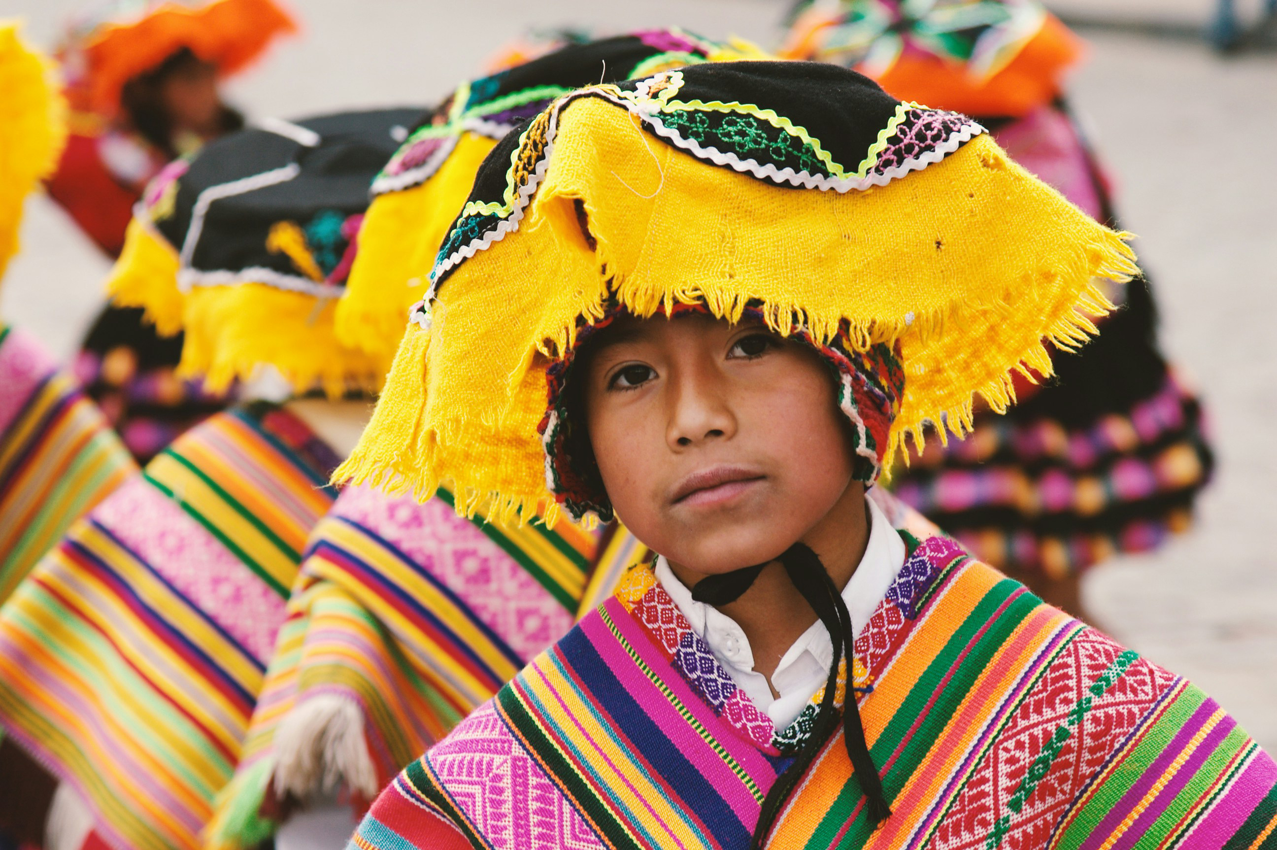 Close-up portrait of a child wearing a bright woven hat and striped poncho, with colorful textiles in background behind them.