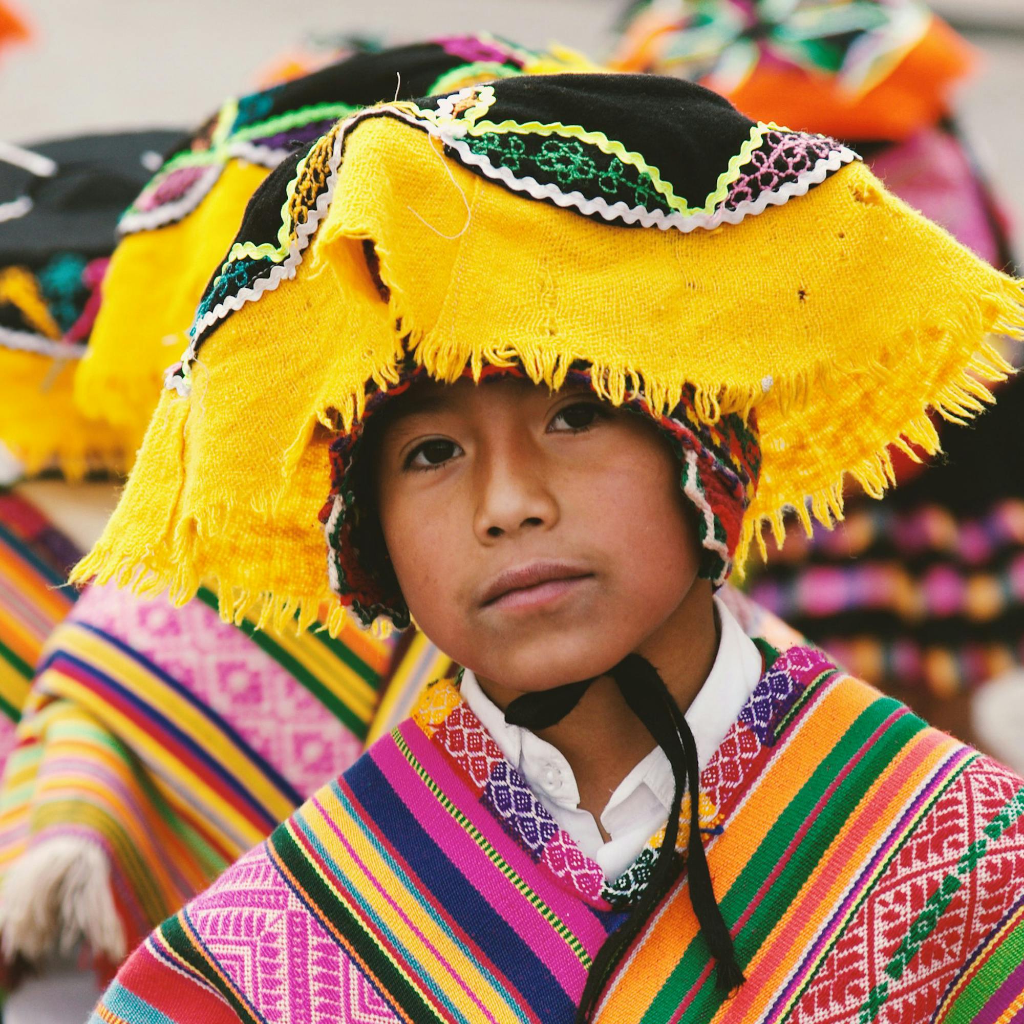 Close-up portrait of a child wearing a bright woven hat and striped poncho, with colorful textiles in background behind them.