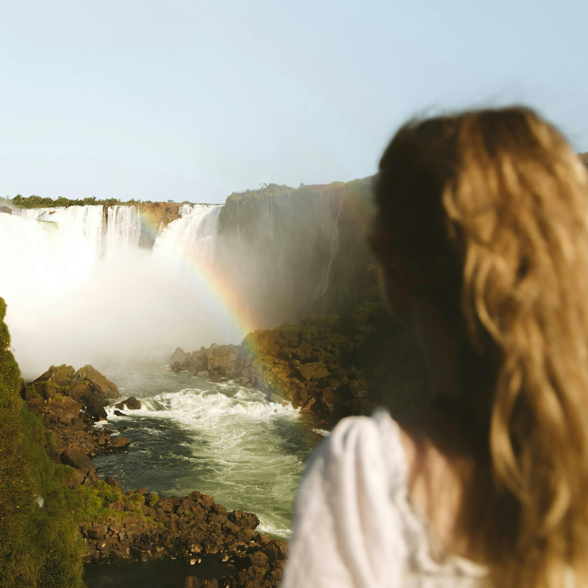 Person stands at a viewpoint overlooking roaring waterfalls and mist, with river rapids and forested cliffs beyond in sunlight.