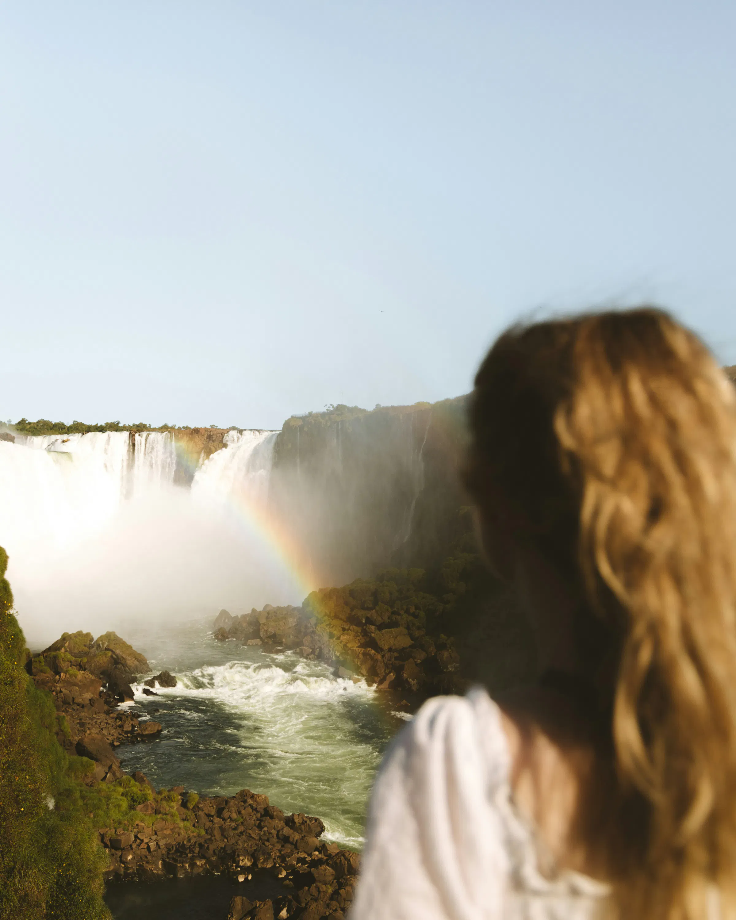 Person stands at a viewpoint overlooking roaring waterfalls and mist, with river rapids and forested cliffs beyond in sunlight.