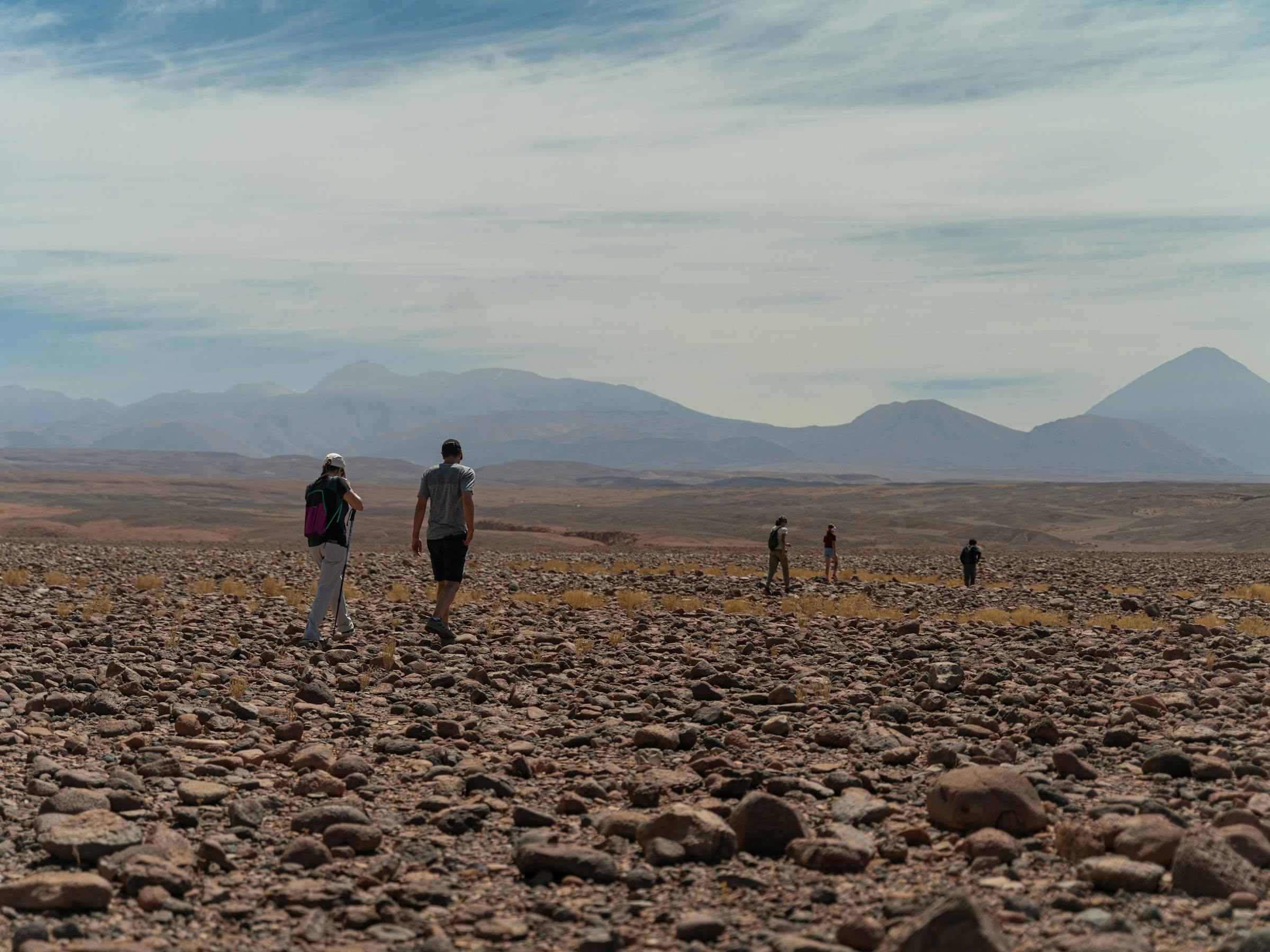 Group of people walk across a rocky desert plain, with low hills and a hazy horizon beneath pale clouds far off.