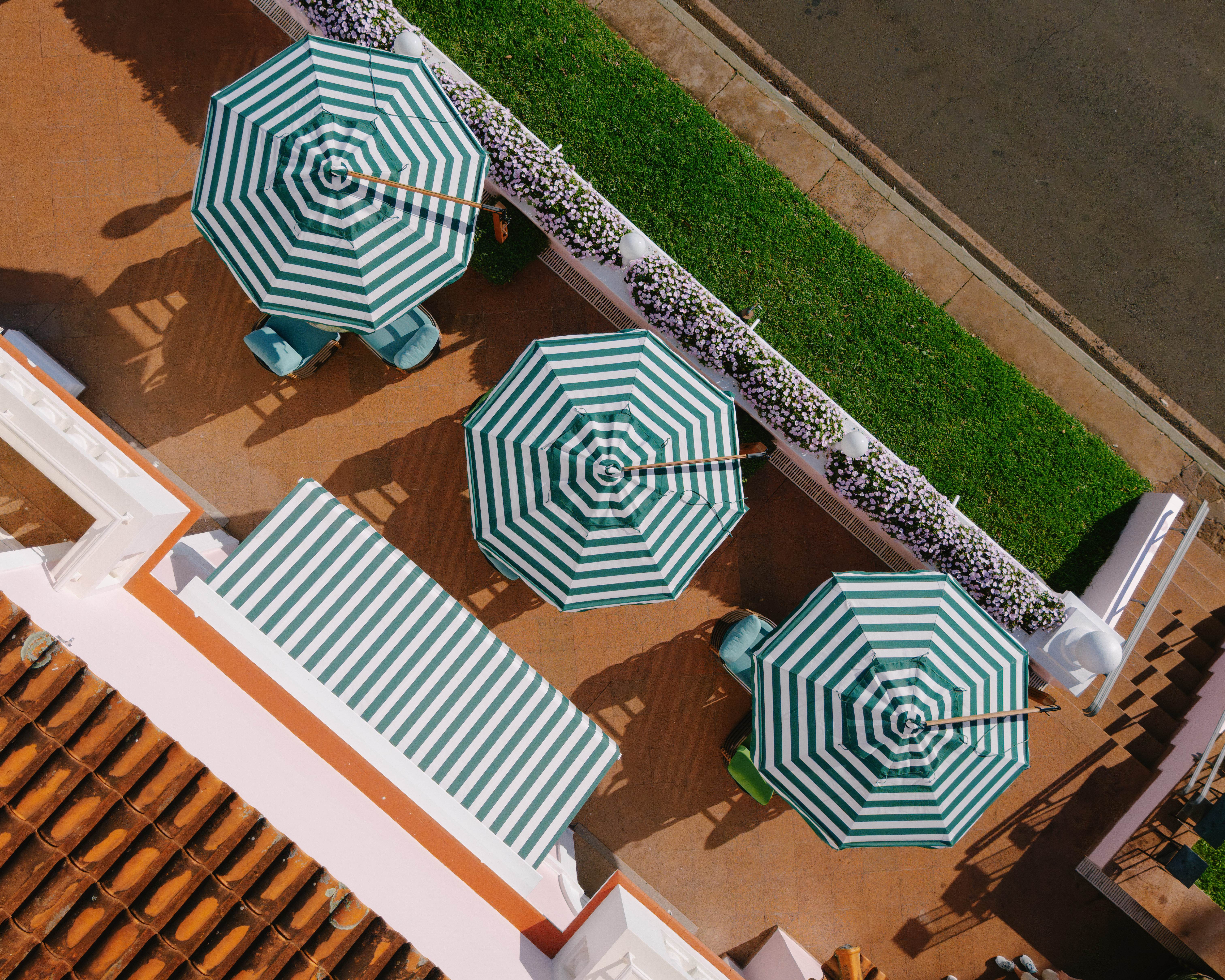 Aerial view of striped umbrellas and loungers on a tan pool deck beside a strip of grass and a paved road.
