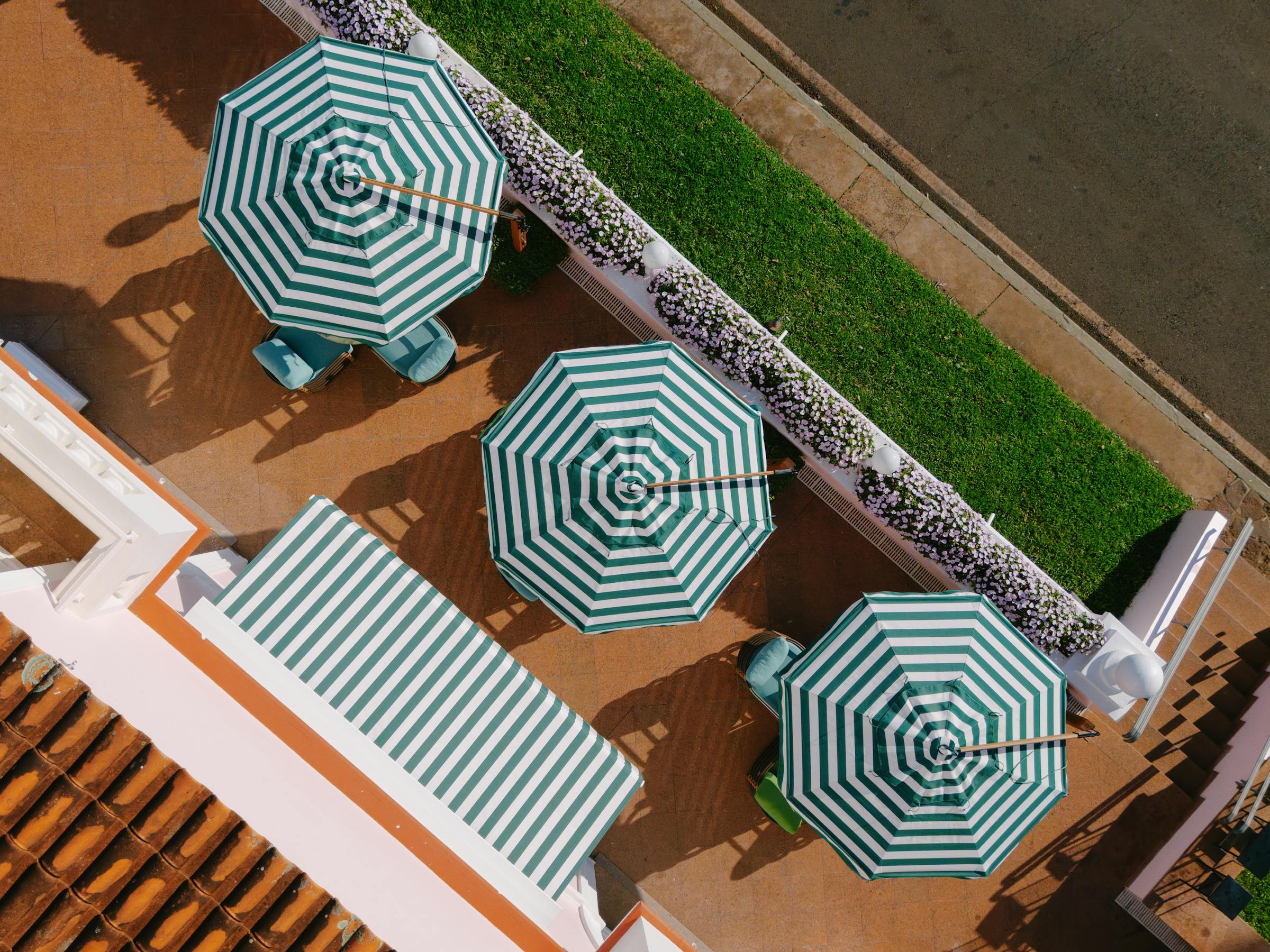 Aerial view of striped umbrellas and loungers on a tan pool deck beside a strip of grass and a paved road.