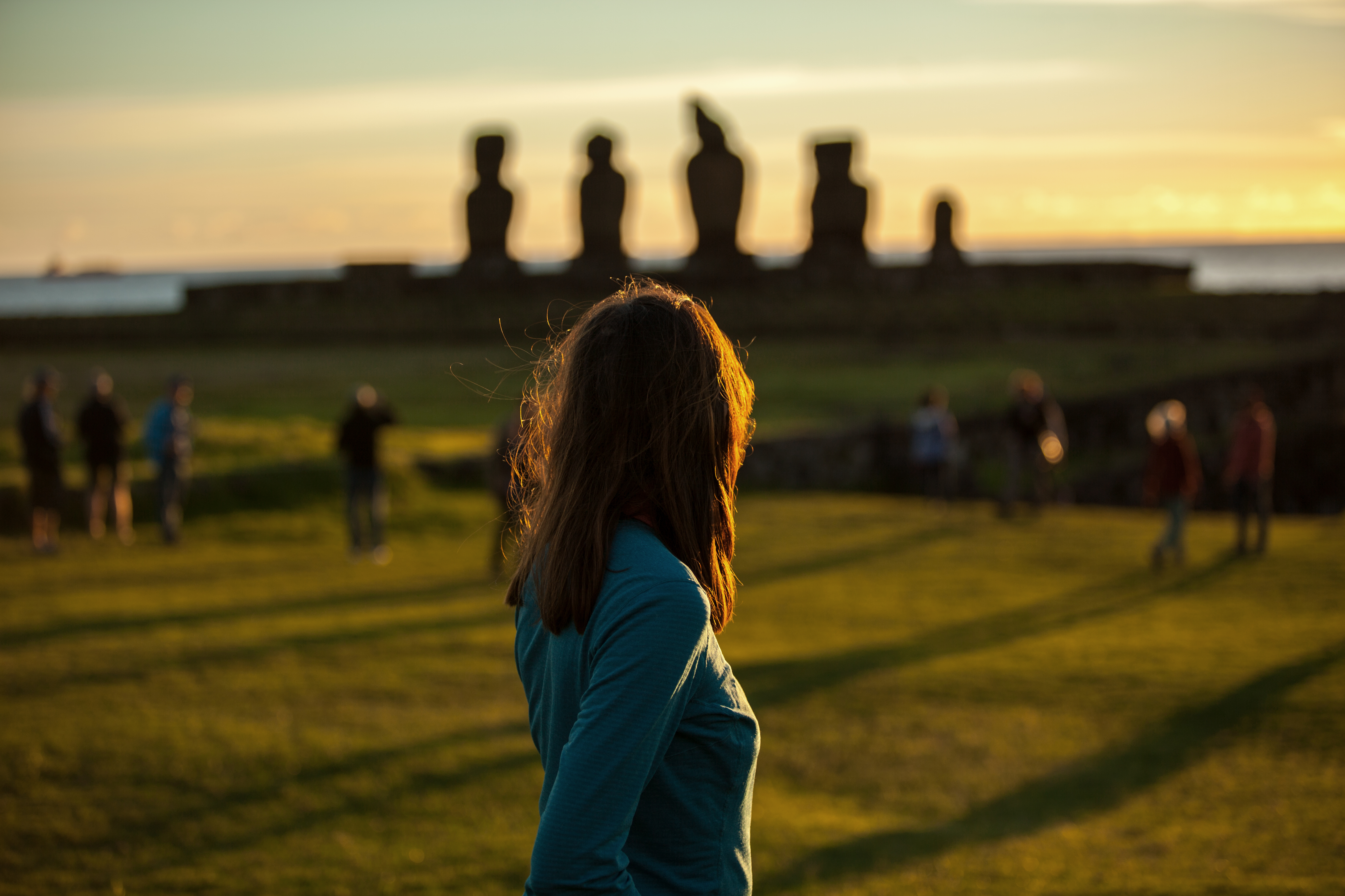Person stands in grassy field looking toward moai statues at sunset, with long shadows and glowing sky behind them quietly.