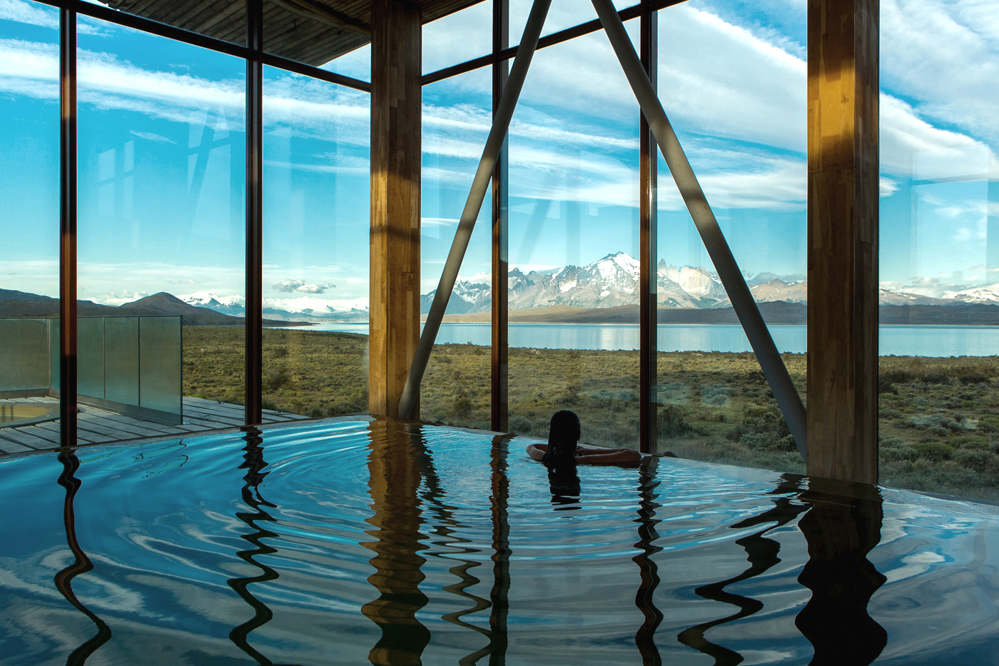 Indoor pool reflects window light, with wooden beams and a panoramic view of water and mountains outside beyond the glass.