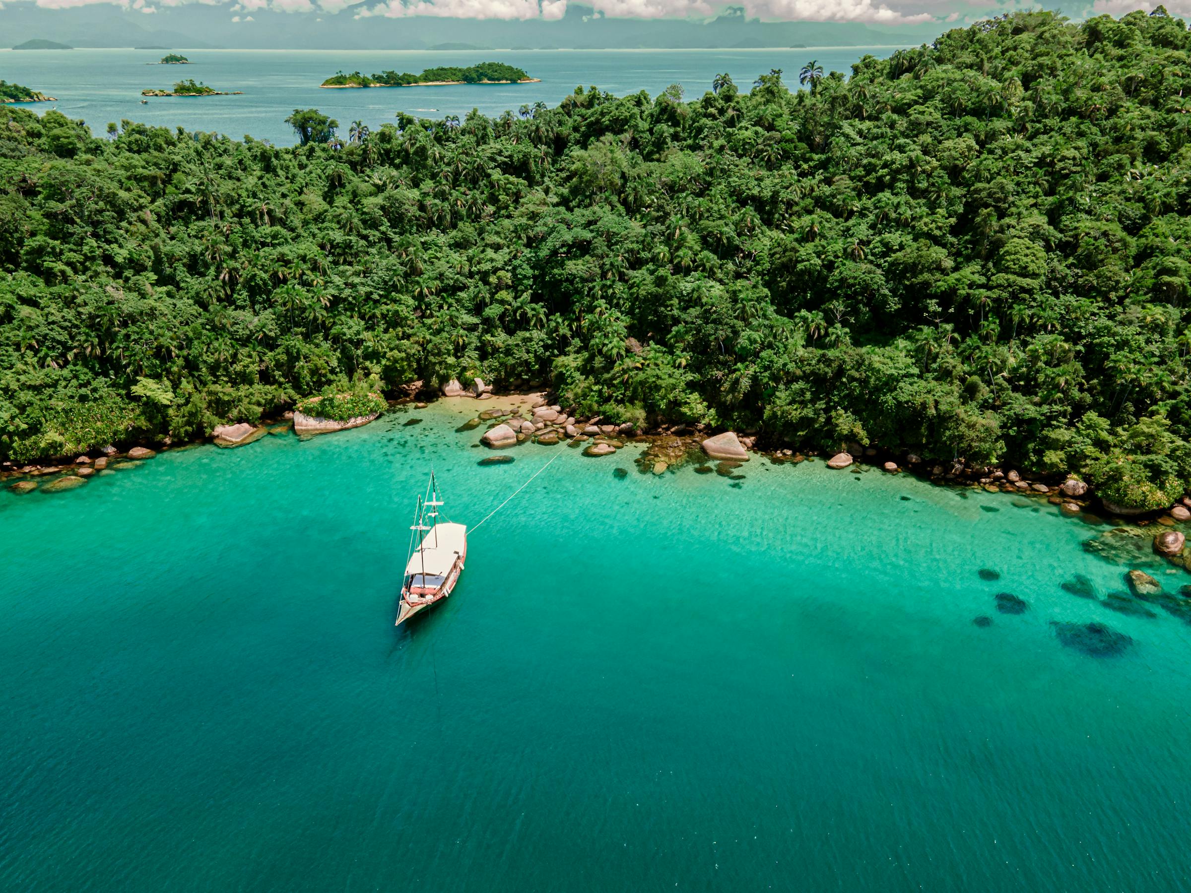 Small boat drifts in a turquoise bay beside dense green shoreline, with shallow sandbars visible beneath clear water nearby.