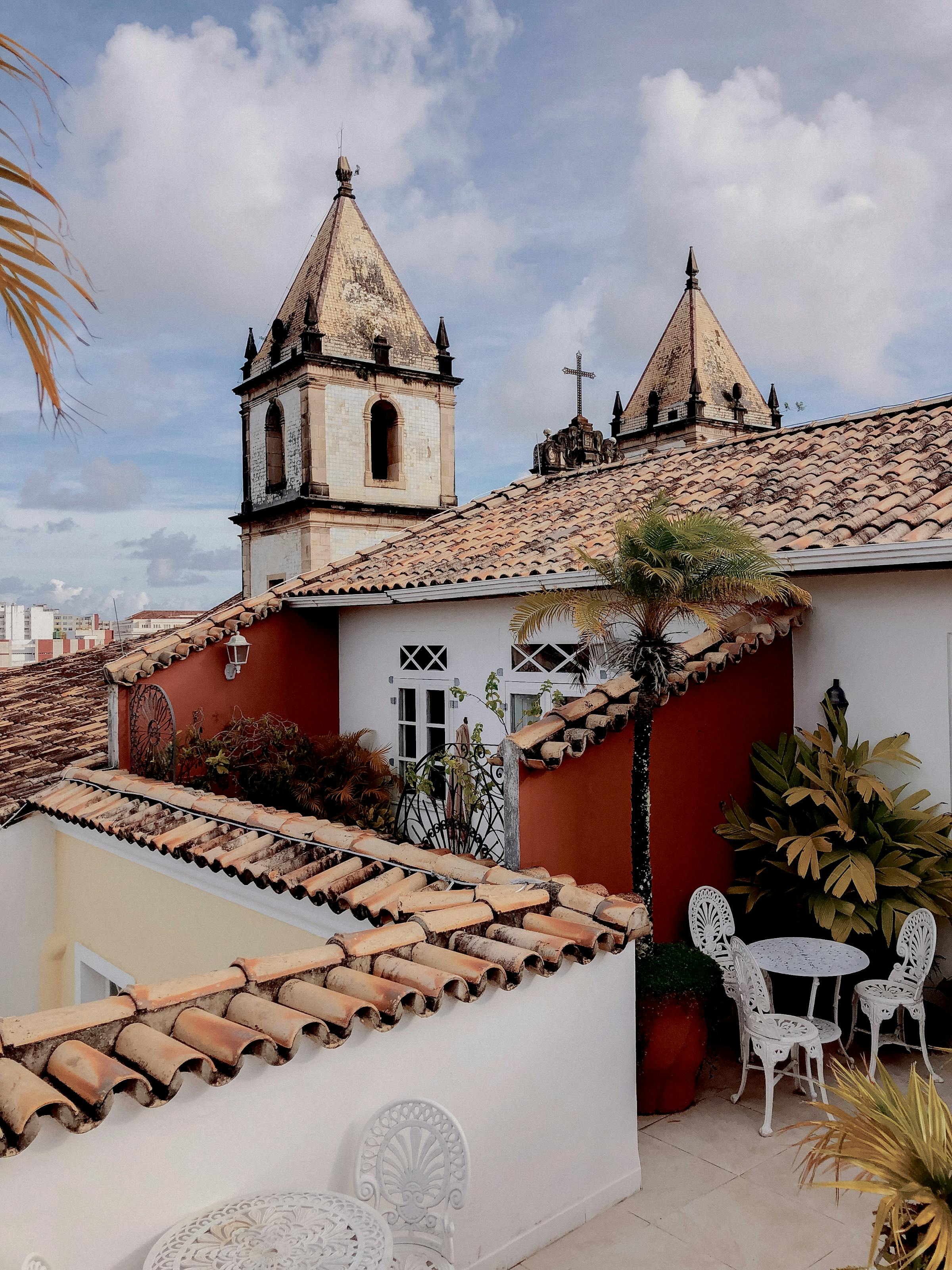 Rooftops and balconies frame a view of a historic church tower, under a soft cloudy sky above the city below.