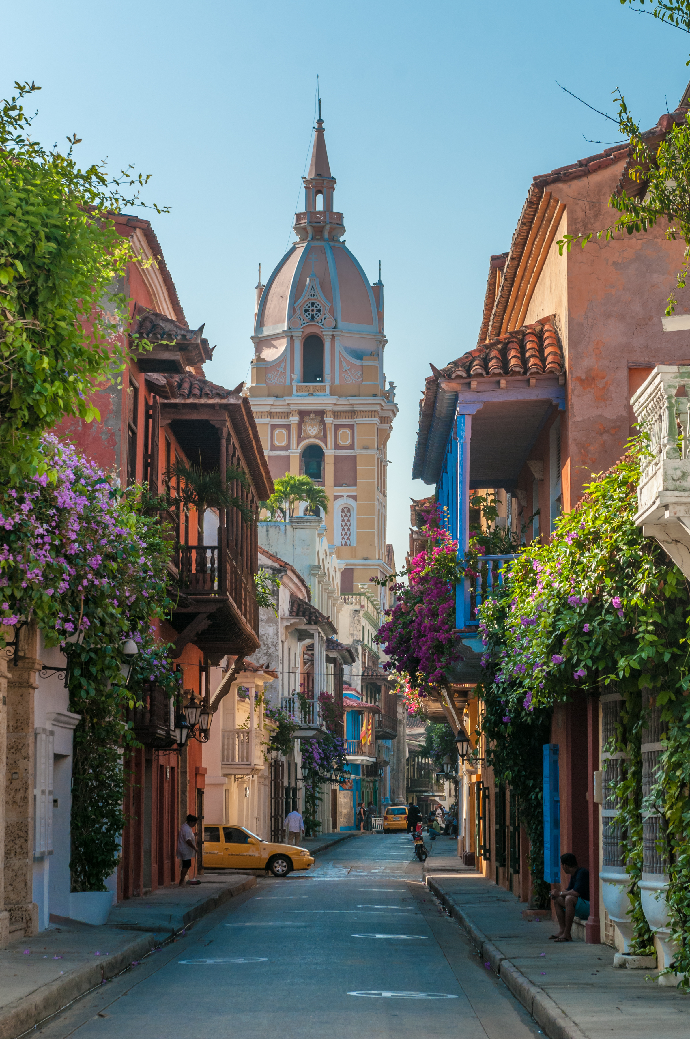 Flower-draped balconies line a narrow cobblestone street leading to a domed church tower in bright sunshine.