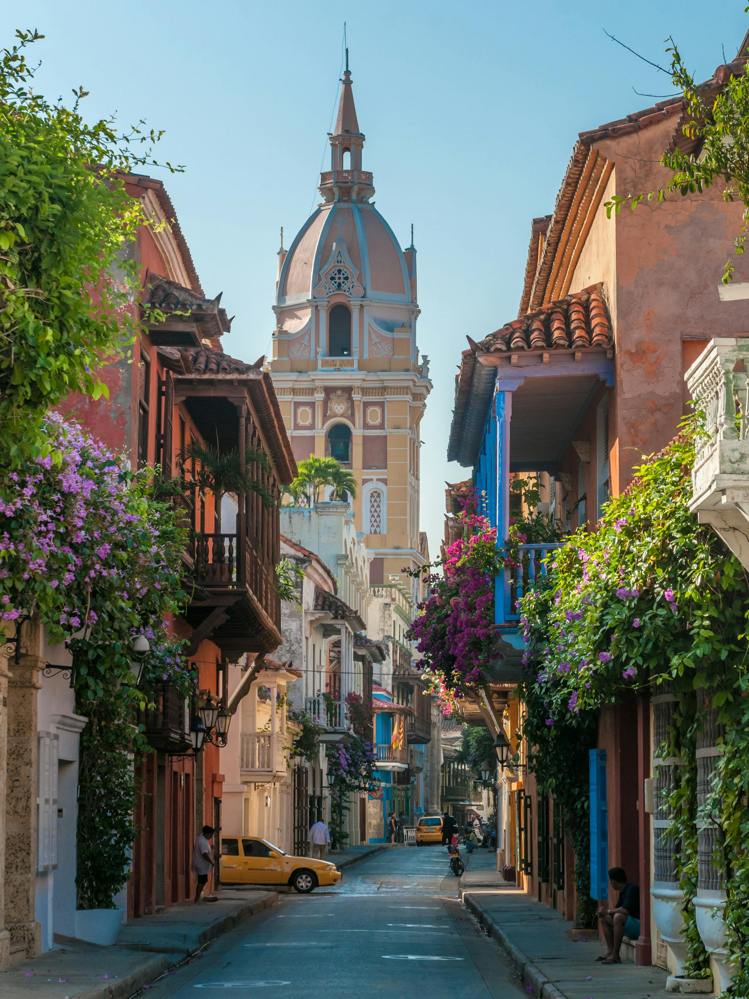 Flower-draped balconies line a narrow cobblestone street leading to a domed church tower in bright sunshine.
