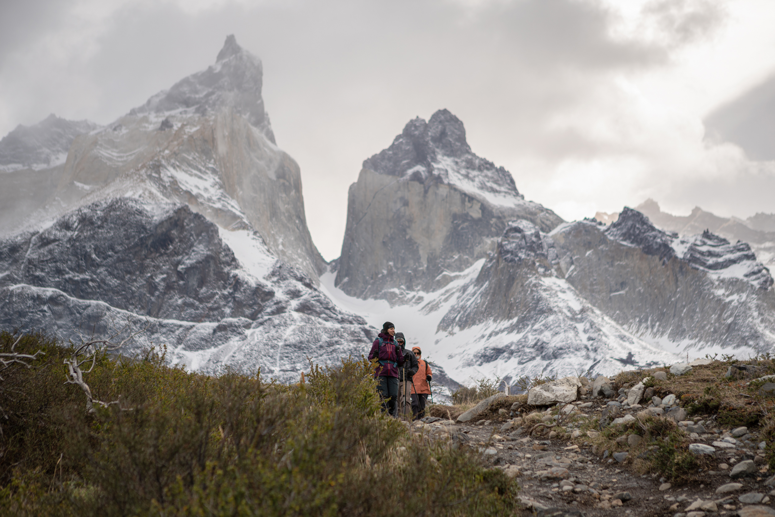 A hiker follows a path toward twin granite peaks, with shrubs in foreground and a cloudy sky overhead ahead.