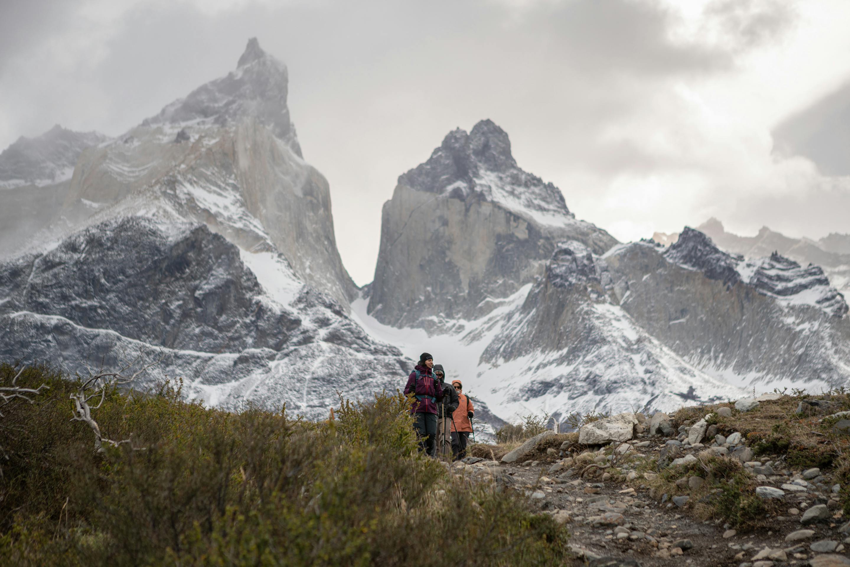 A hiker follows a path toward twin granite peaks, with shrubs in foreground and a cloudy sky overhead ahead.