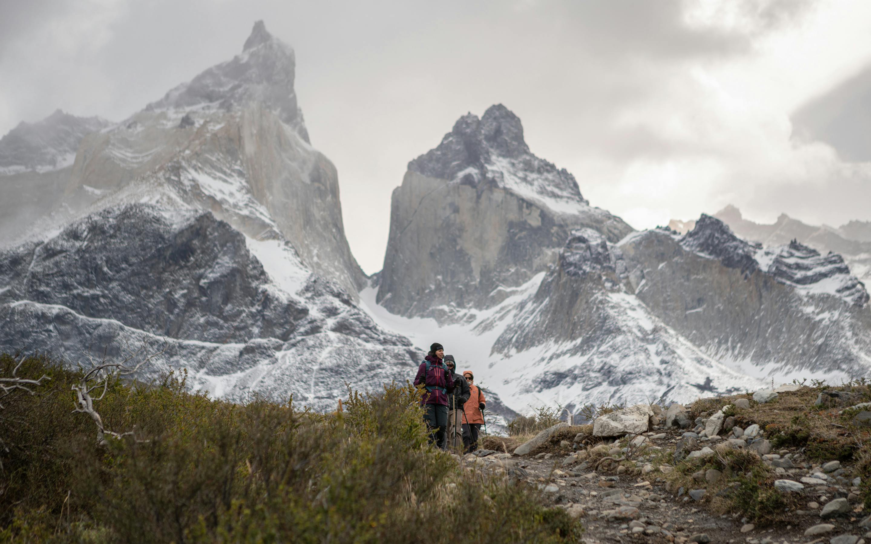 A hiker follows a path toward twin granite peaks, with shrubs in foreground and a cloudy sky overhead ahead.