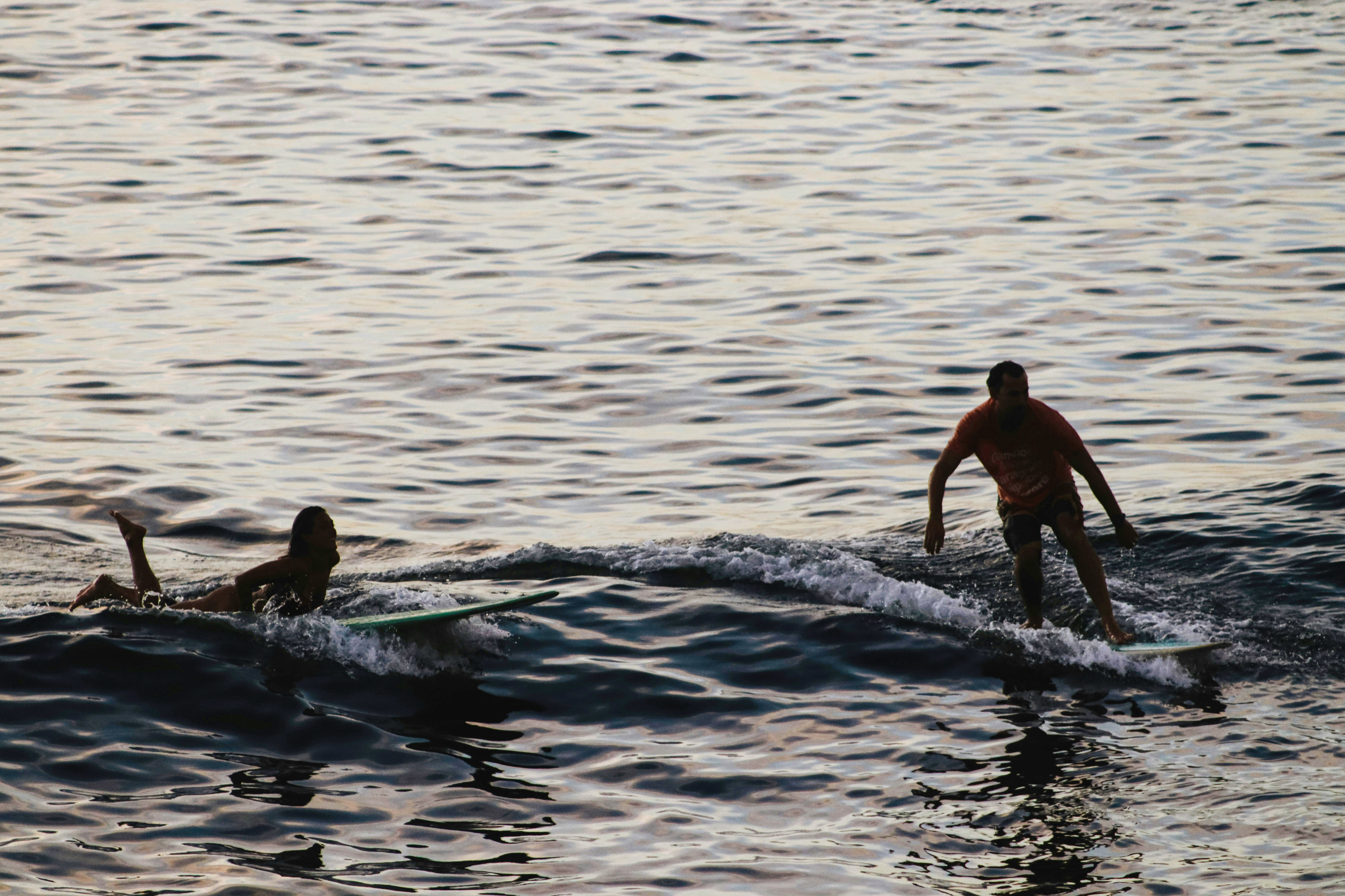 Two surfers ride small waves at dusk, one standing on a board while another paddles nearby on rippled water.