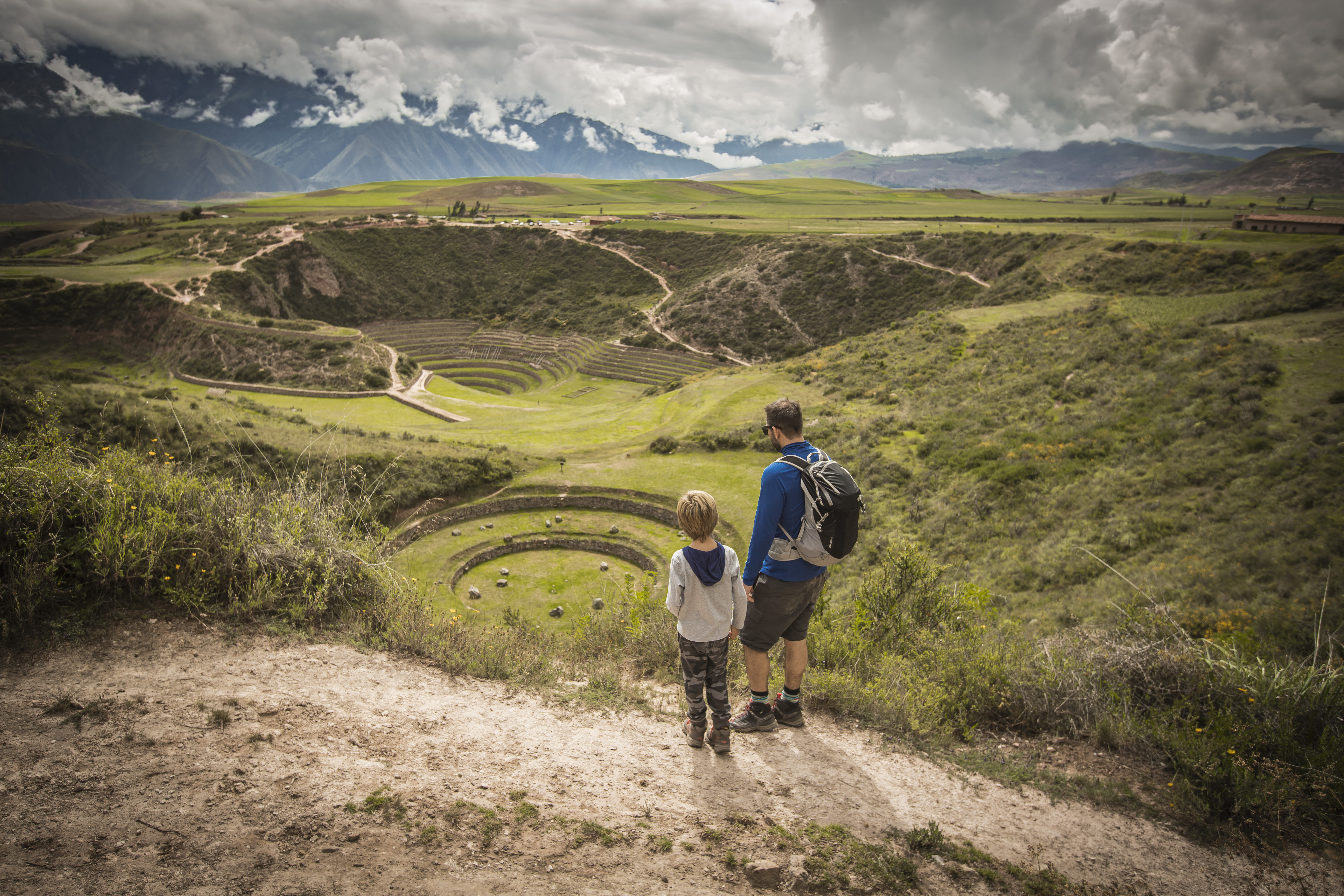 Two hikers walk along a dirt path through a wide green valley, with layered hills and clouds overhead together slowly.