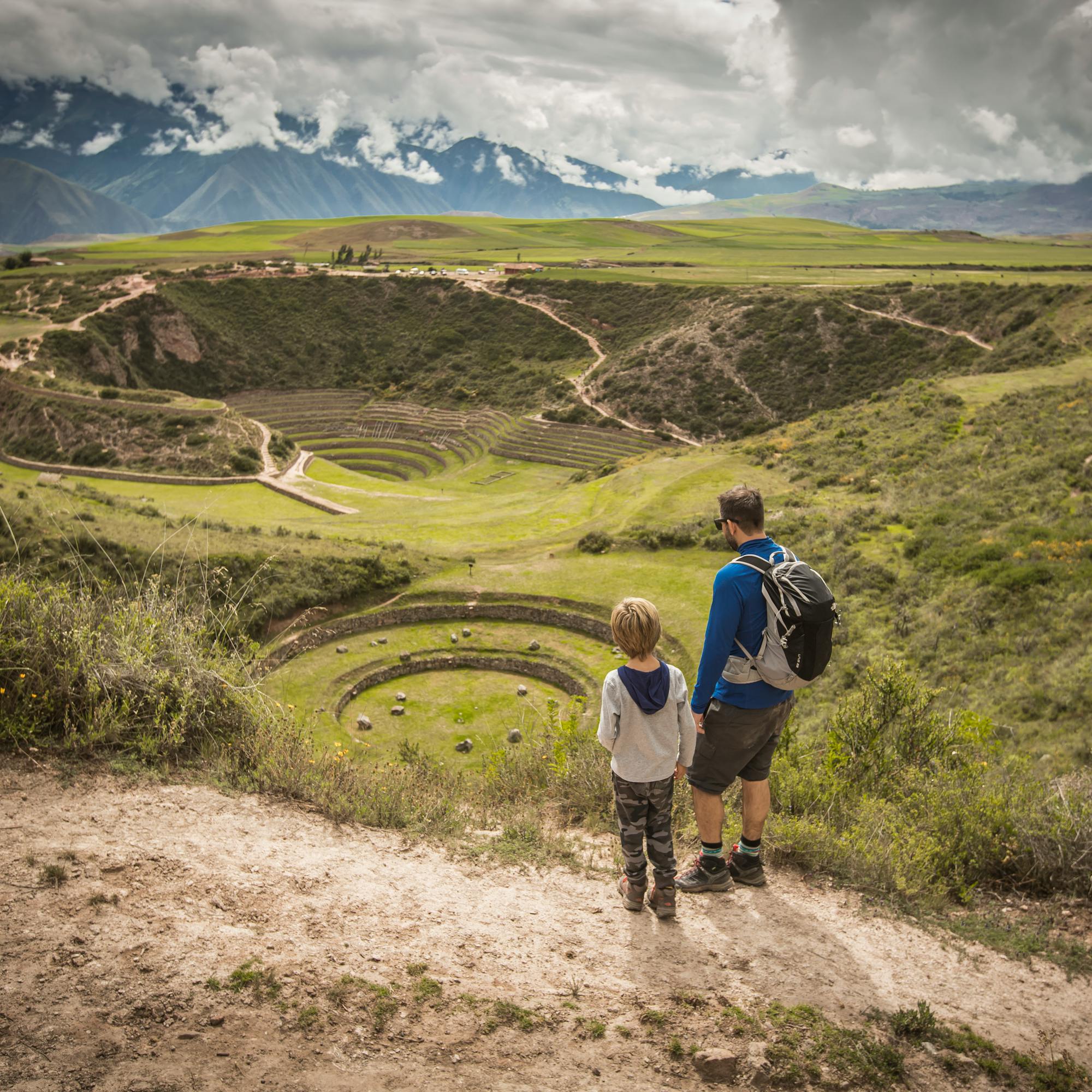 Two hikers walk along a dirt path through a wide green valley, with layered hills and clouds overhead together slowly.
