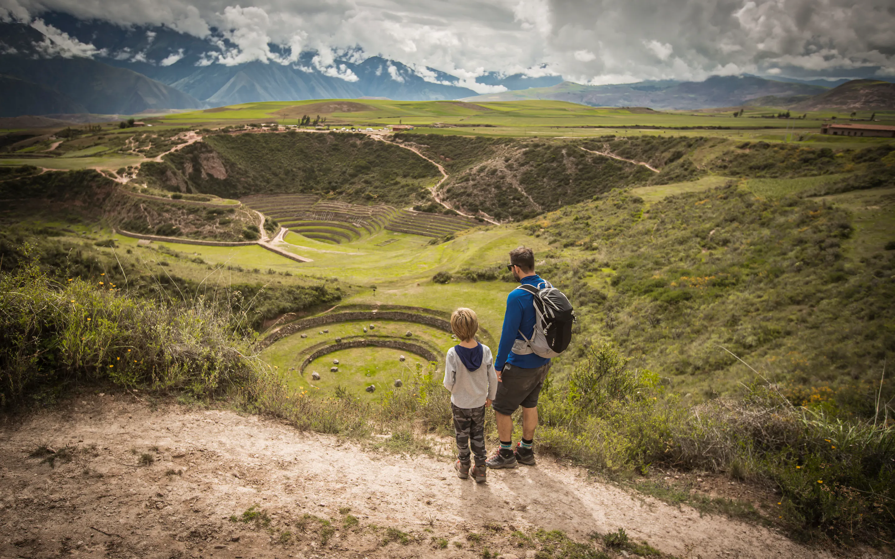 Two hikers walk along a dirt path through a wide green valley, with layered hills and clouds overhead together slowly.