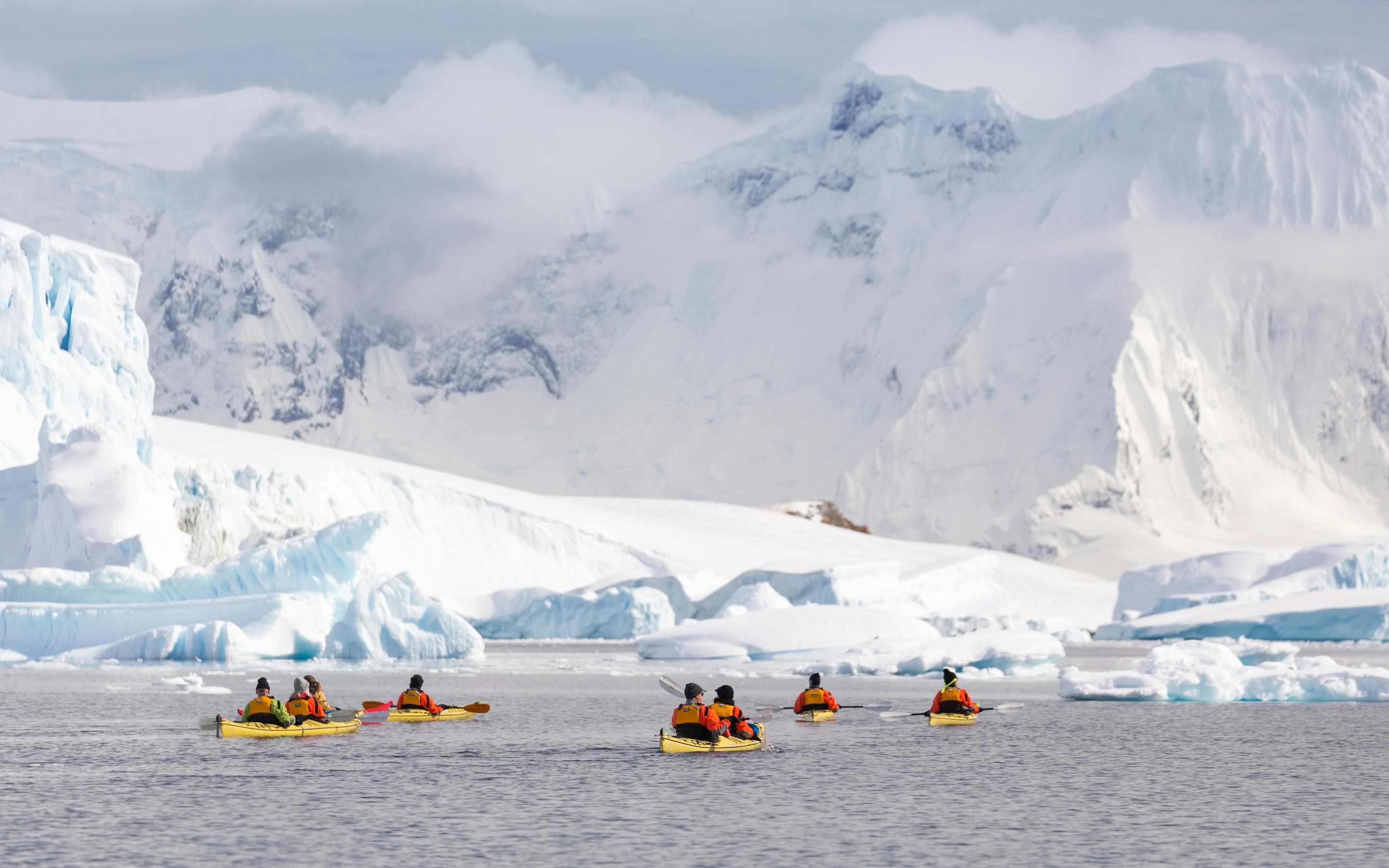 Bright kayaks float on icy water near glaciers, with snow-covered peaks and mist drifting over the bay around them.