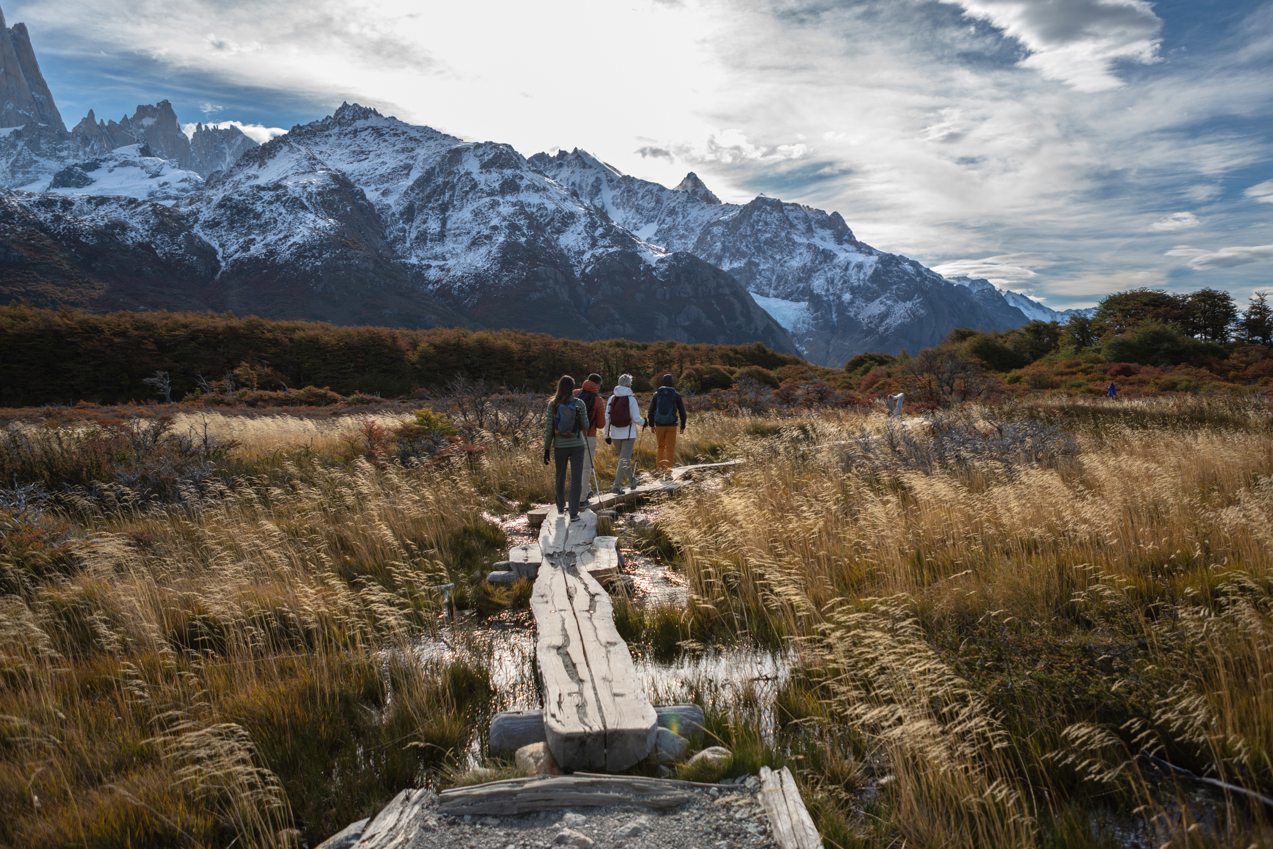 Hikers walk along a wooden trail through golden grassland, with snowcapped mountains and dramatic clouds ahead in cool wind .