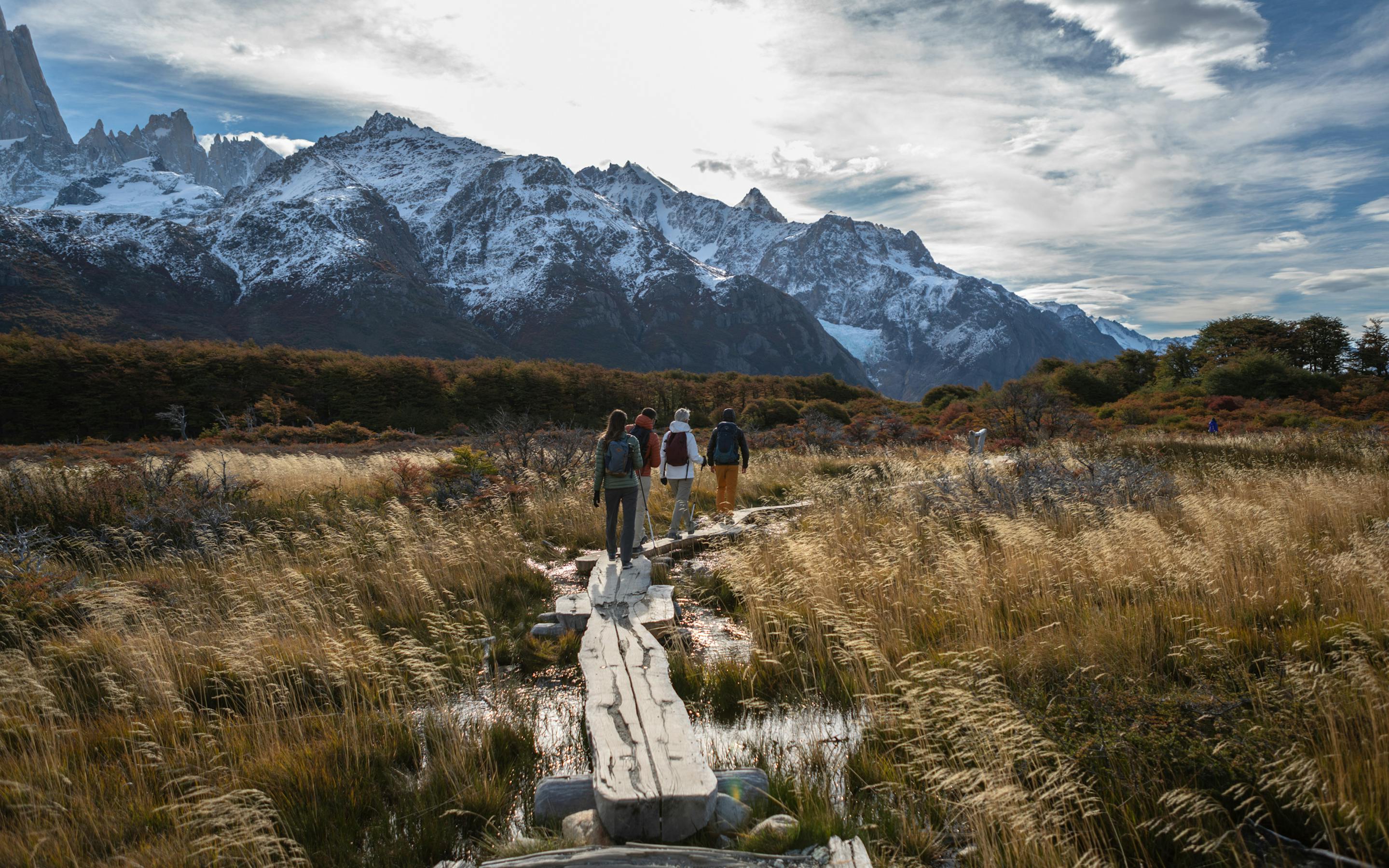 Hikers walk along a wooden trail through golden grassland, with snowcapped mountains and dramatic clouds ahead in cool wind .