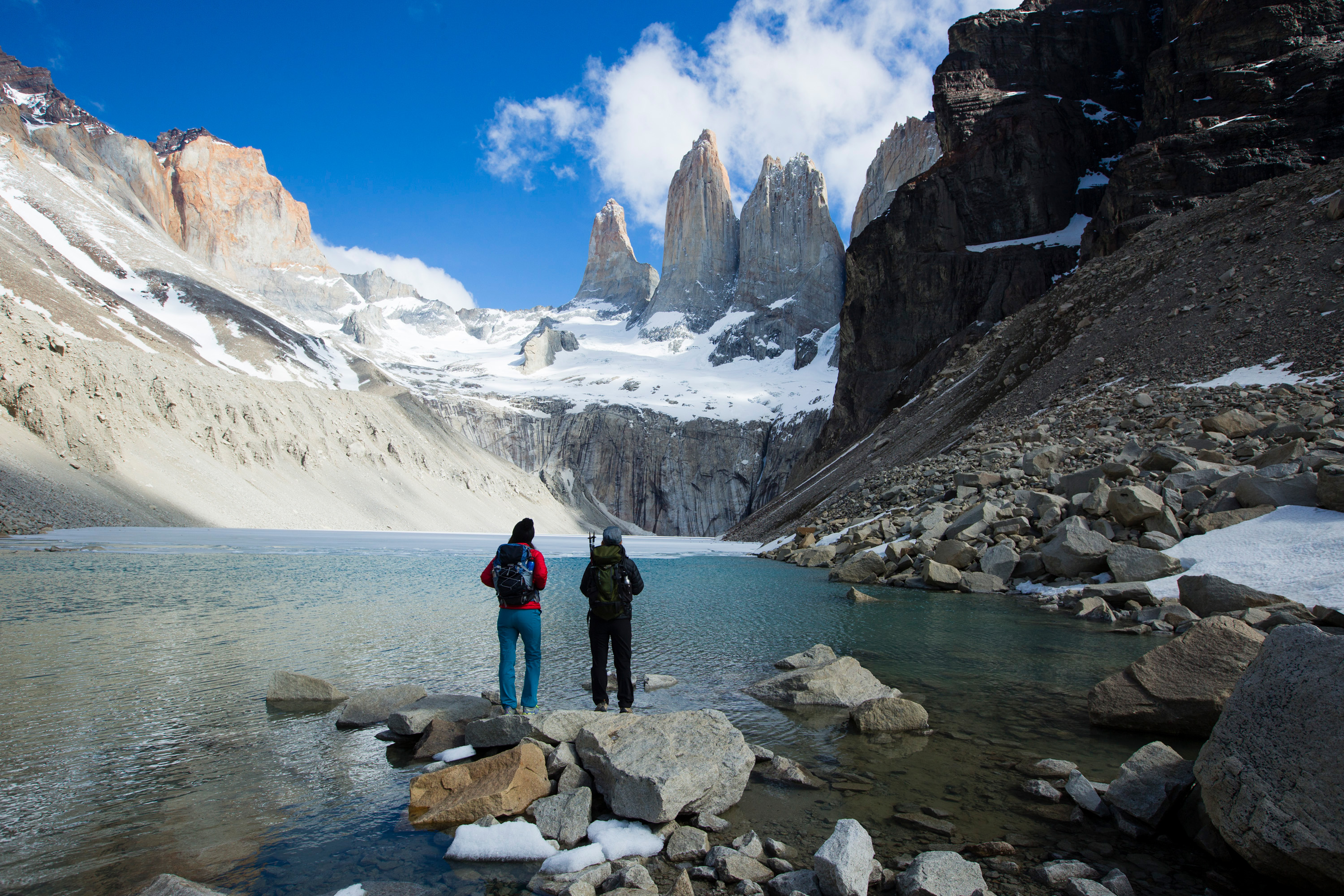 Hikers stand beside a turquoise glacial lake surrounded by jagged peaks, with snow patches and clear reflections nearby.