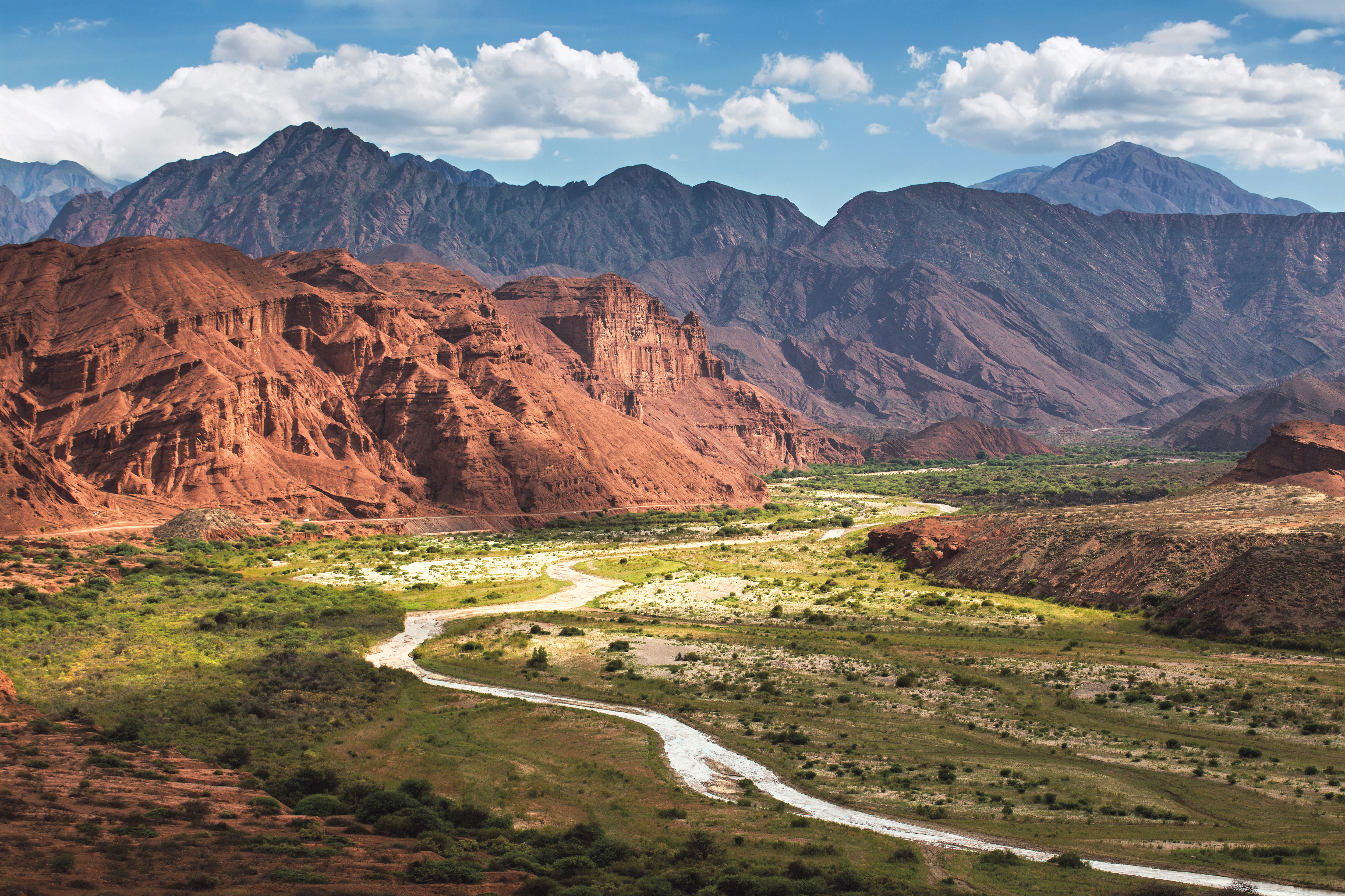 Winding river cuts through a broad red canyon, with layered mountains and blue haze stretching into distance behind it all.