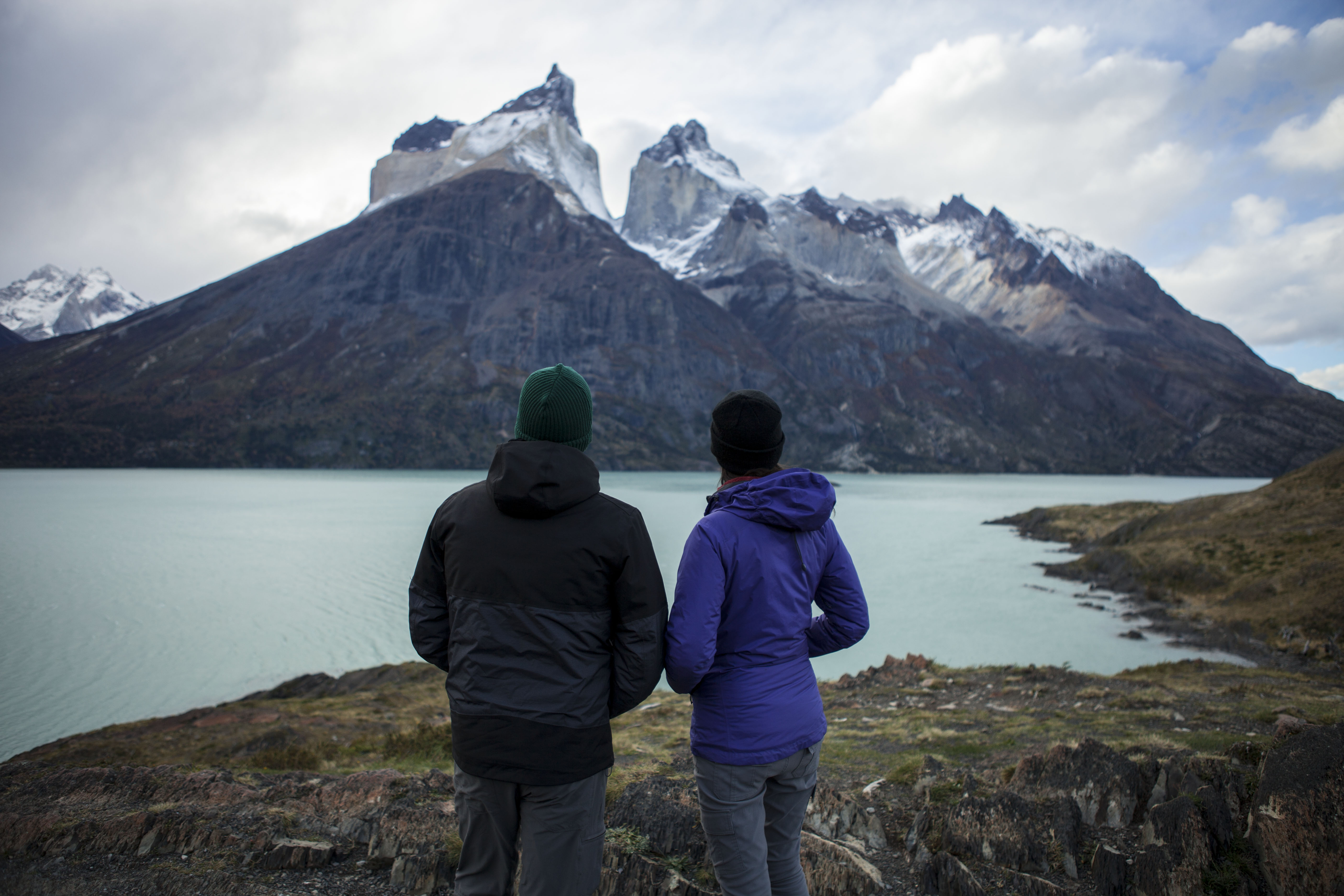 Two figures in jackets stand at a lakeside overlook, facing dramatic gray mountains and a calm turquoise bay below them.