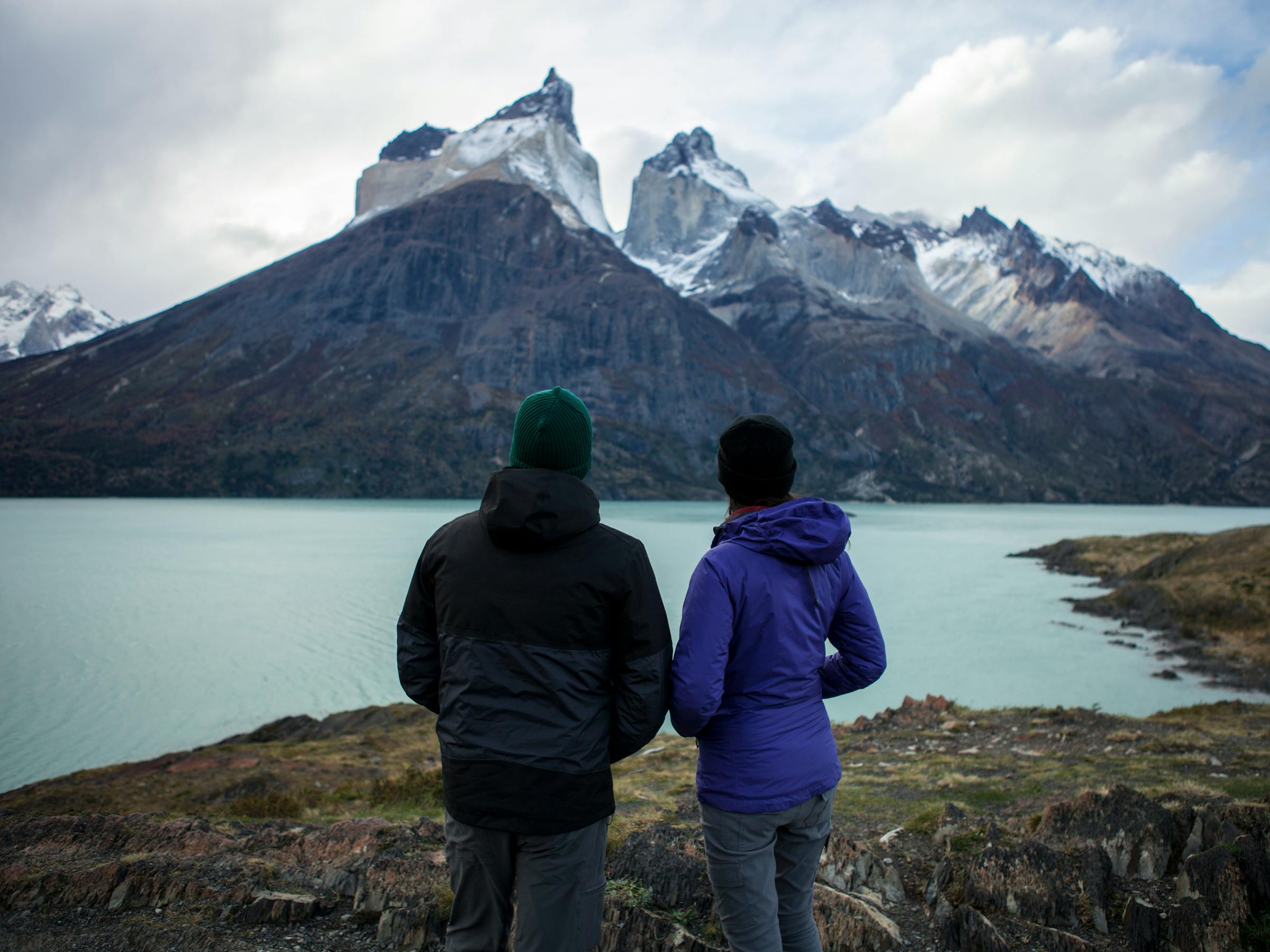 Two figures in jackets stand at a lakeside overlook, facing dramatic gray mountains and a calm turquoise bay below them.