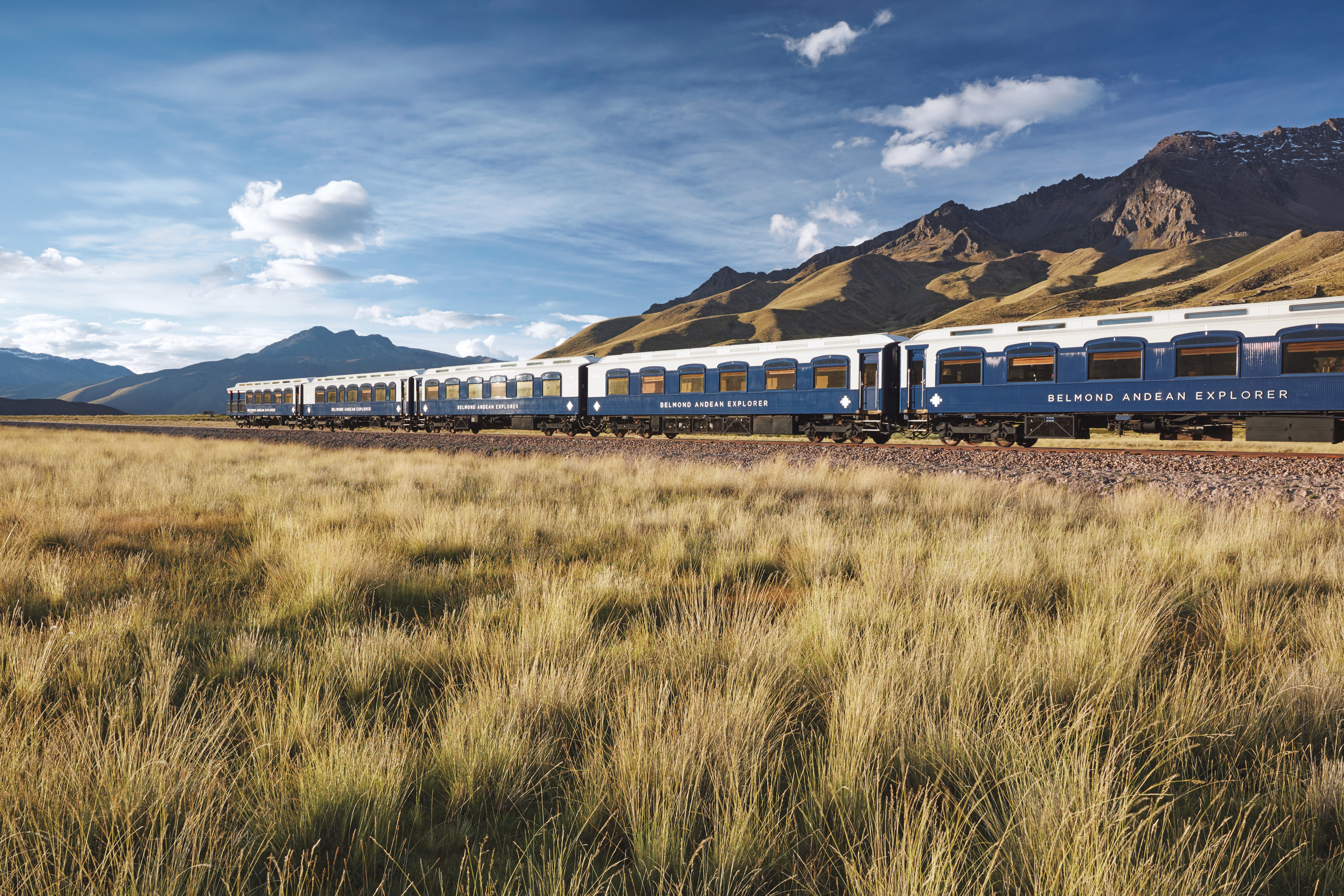 A blue-and-white train travels across golden grasslands, with broad Andean mountains and scattered clouds in background under bright daylight
