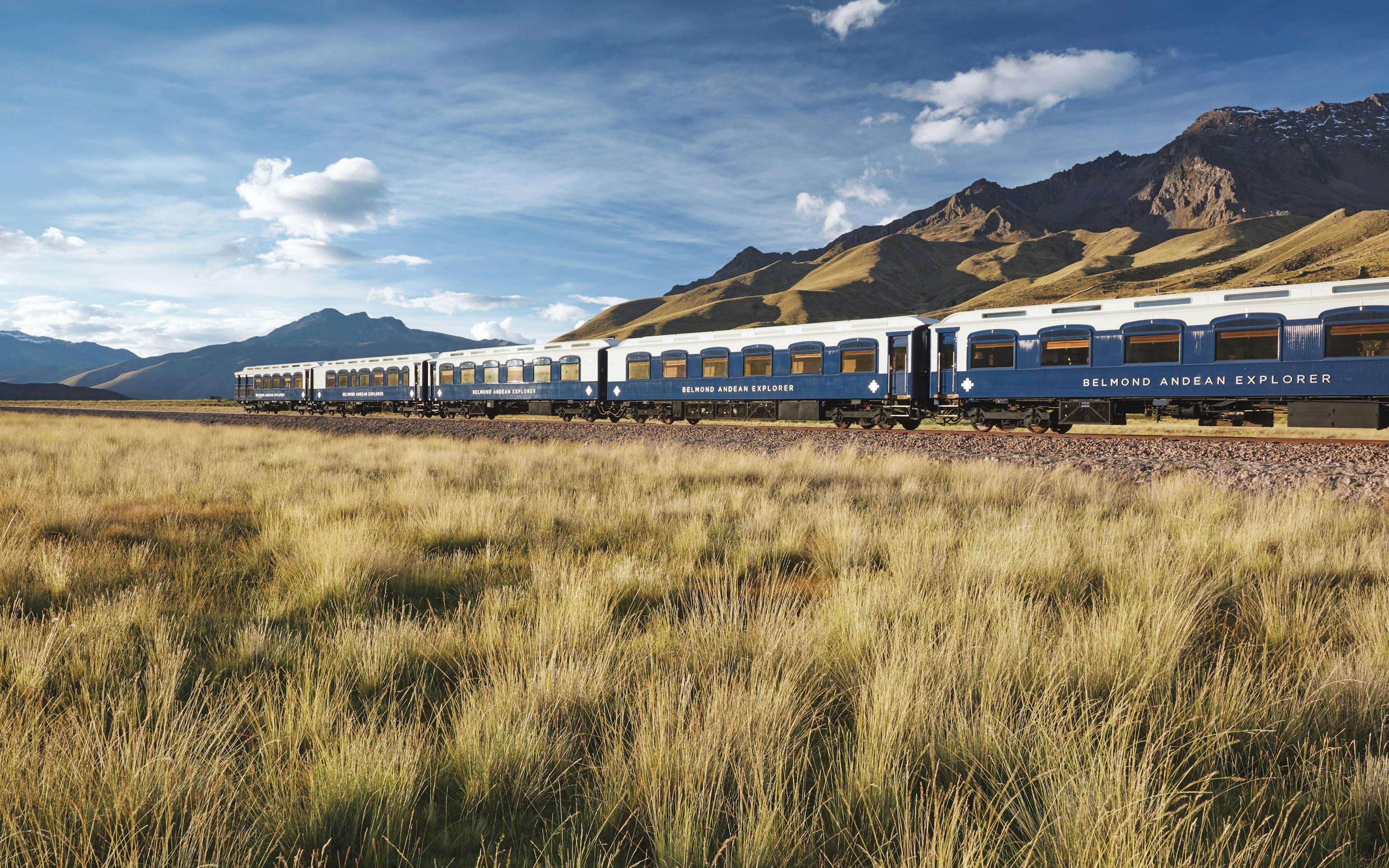 A blue-and-white train travels across golden grasslands, with broad Andean mountains and scattered clouds in background under bright daylight