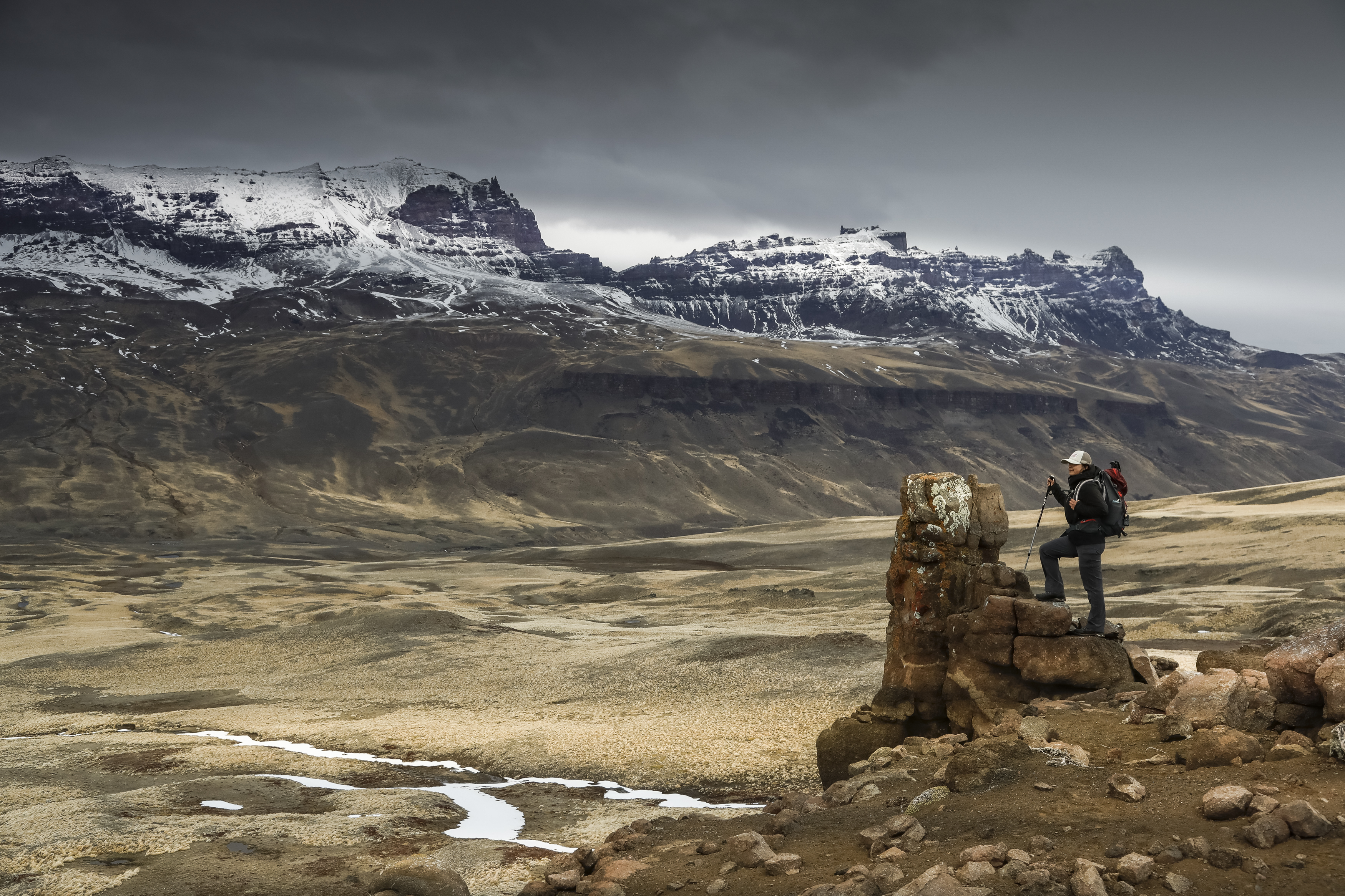 Person stands on a boulder overlooking wide plains and jagged peaks, under gray skies and windy conditions far ahead.