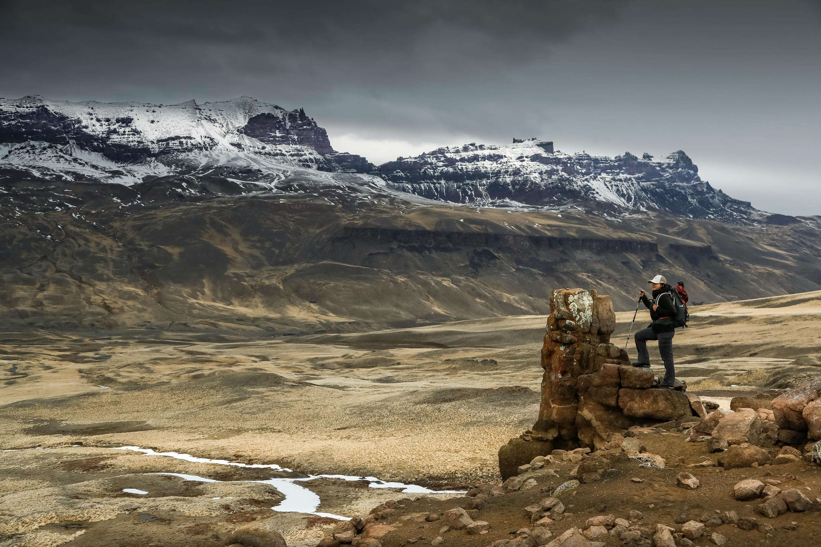 Person stands on a boulder overlooking wide plains and jagged peaks, under gray skies and windy conditions far ahead.
