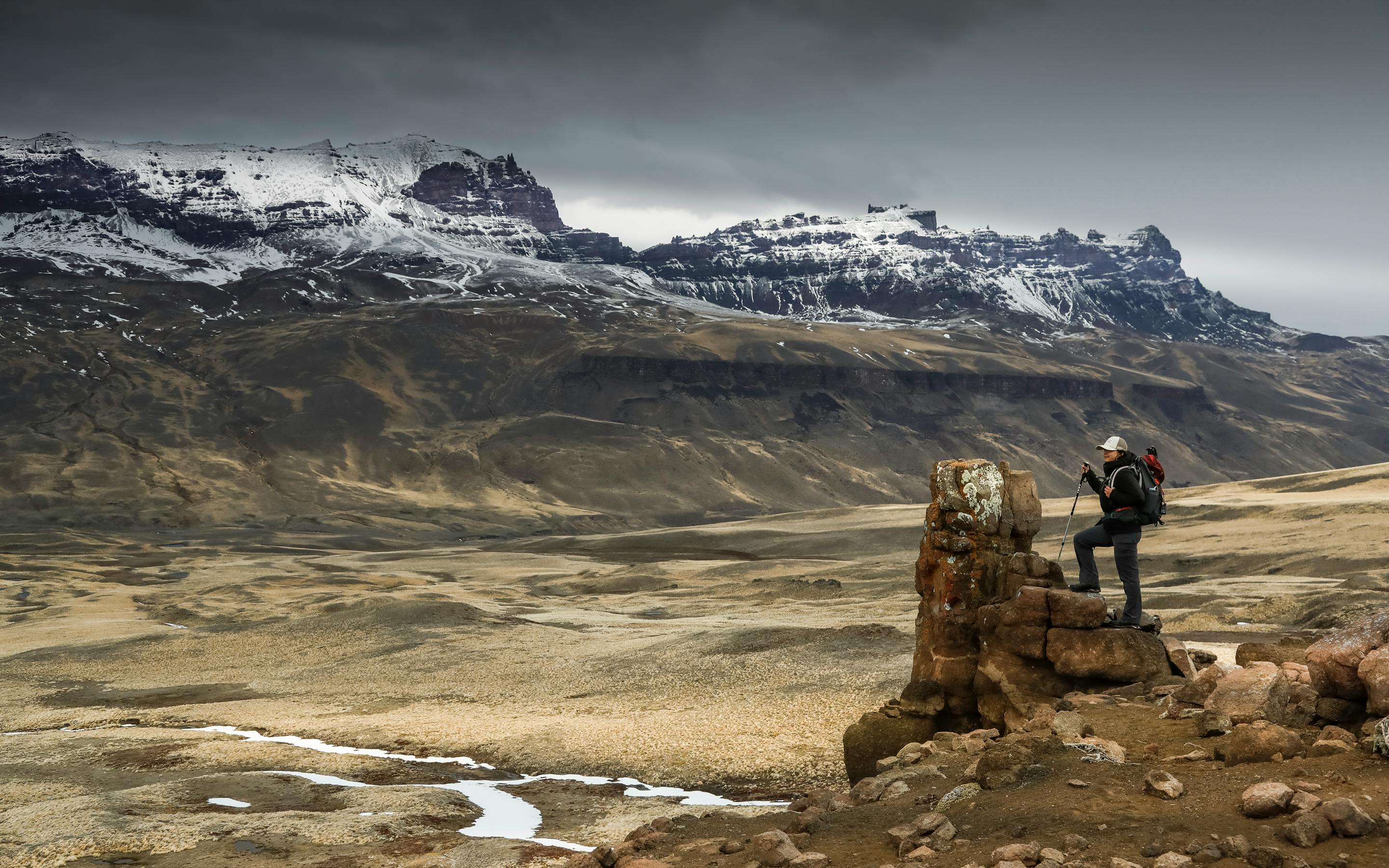 Person stands on a boulder overlooking wide plains and jagged peaks, under gray skies and windy conditions far ahead.