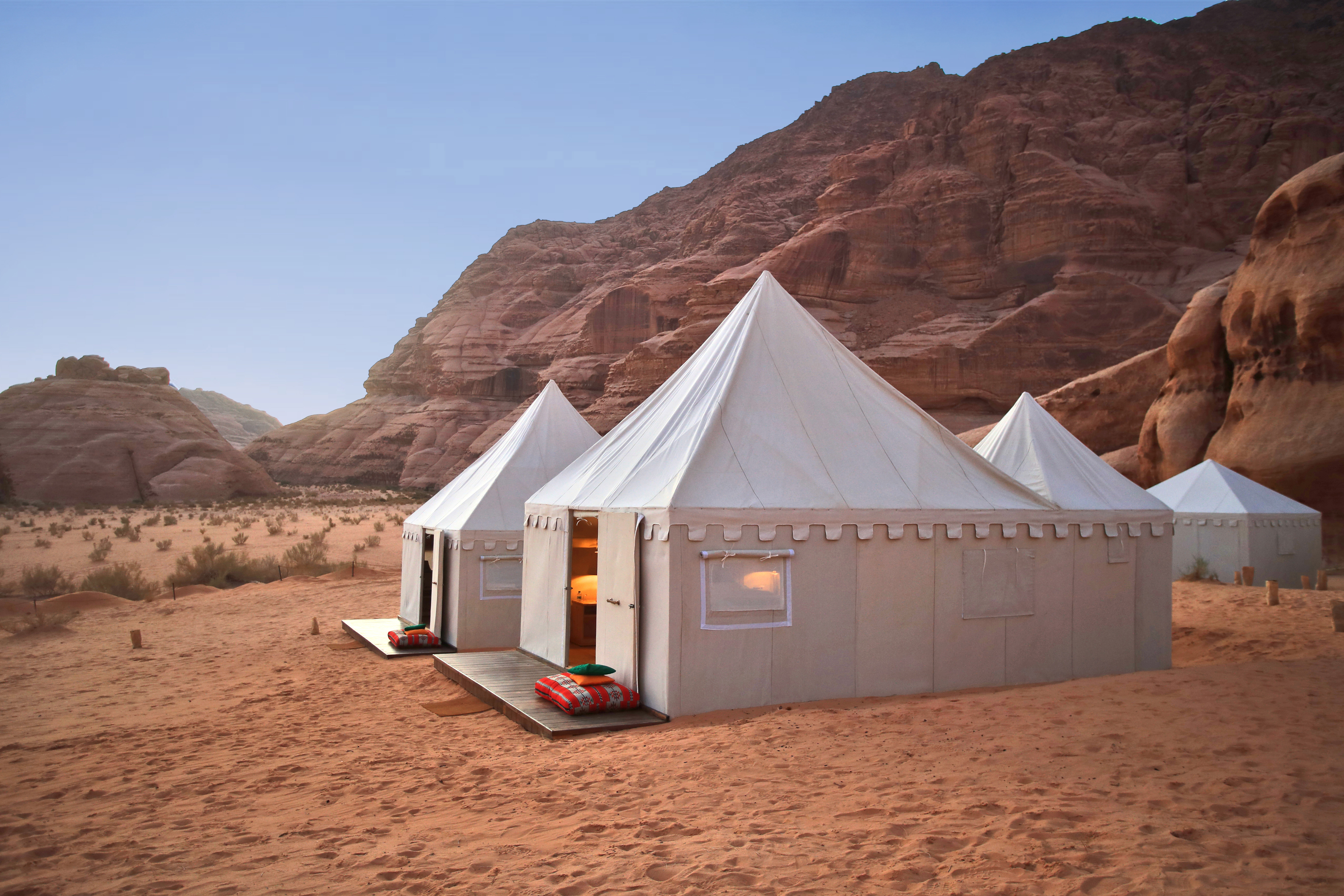 White tents set on sandy ground beside towering red rock cliffs.