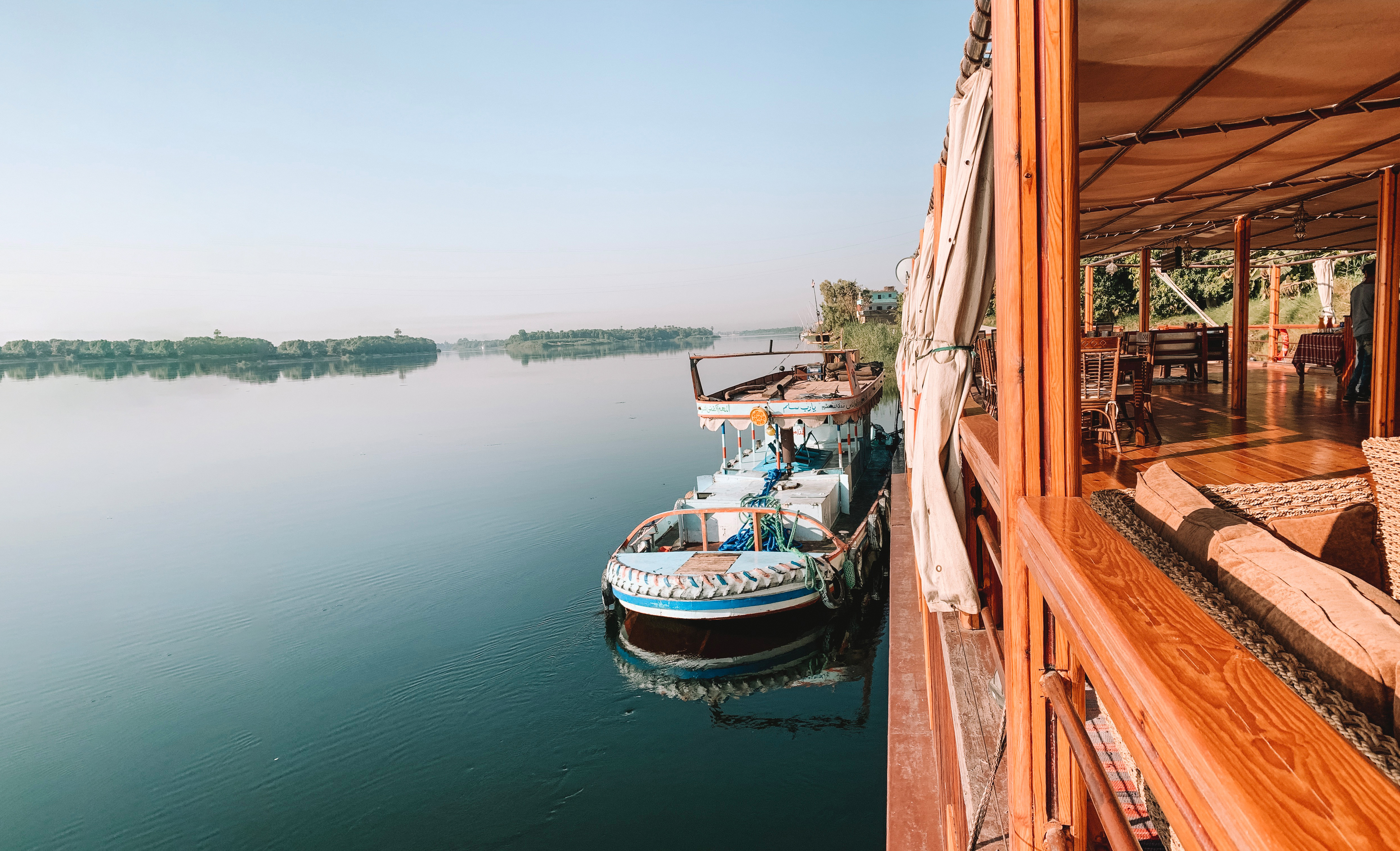 Lounge chairs line a wooden boat deck beside calm blue water with another boat nearby.