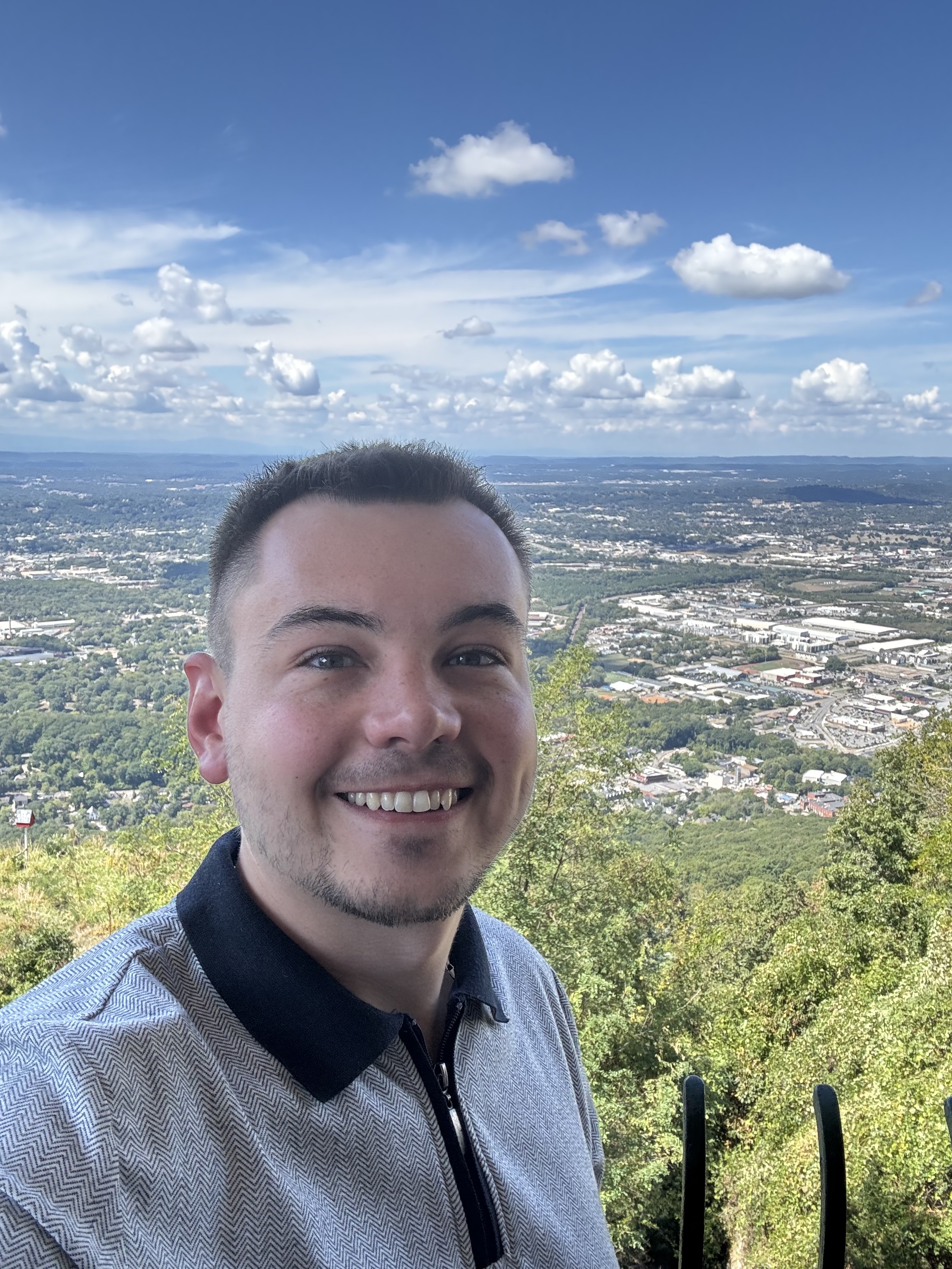 Smiling man standing at a scenic overlook with a wide view of a city, green hills, and a blue sky with scattered clouds in the background.