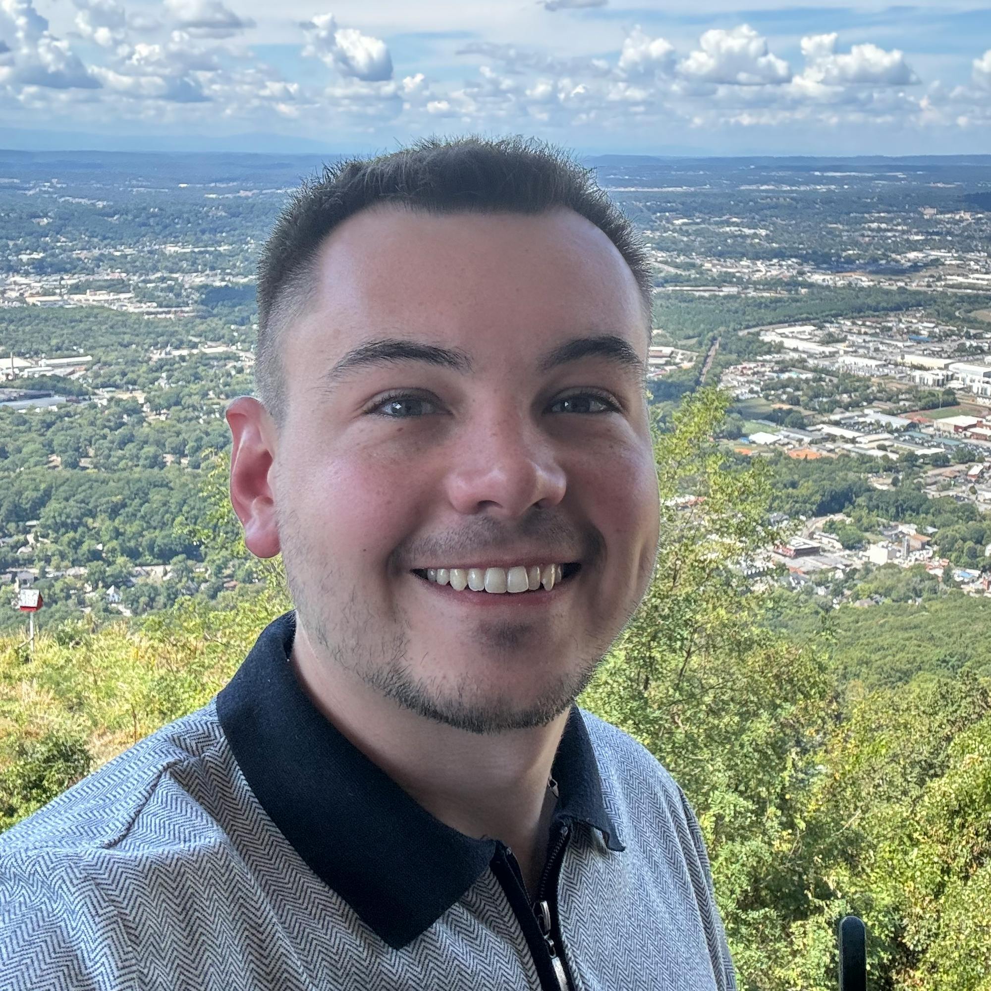 Smiling man standing at a scenic overlook with a wide view of a city, green hills, and a blue sky with scattered clouds in the background.