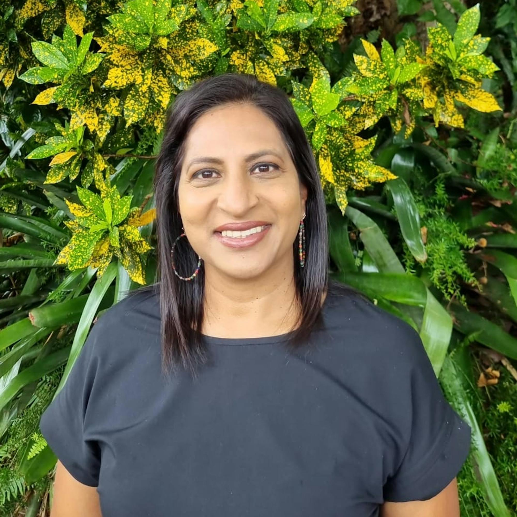 Smiling woman standing outdoors in front of lush green foliage with yellow-speckled leaves, wearing a black top.