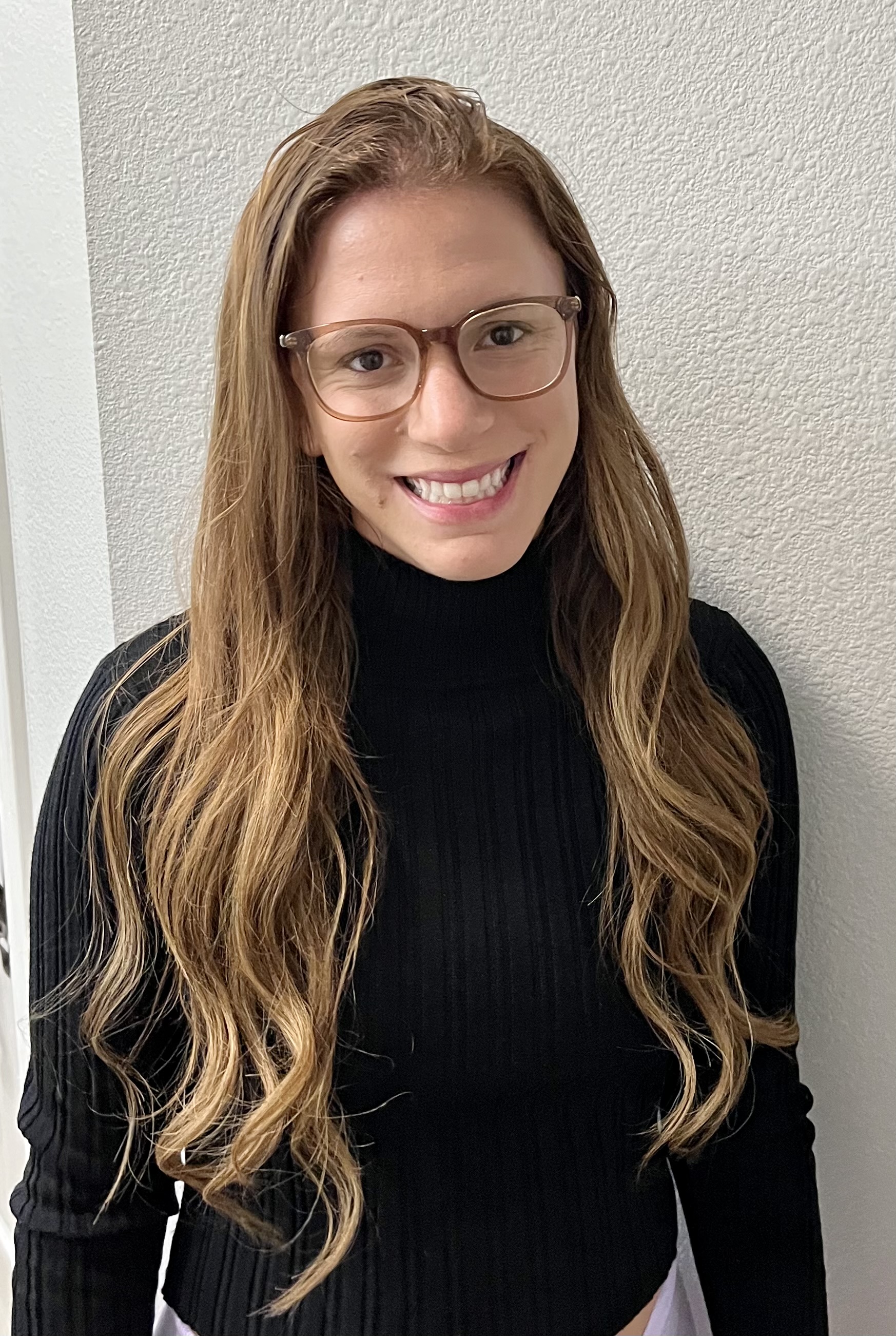 Smiling woman with long light brown hair and glasses, wearing a black long-sleeve top, standing against a light-colored textured wall.