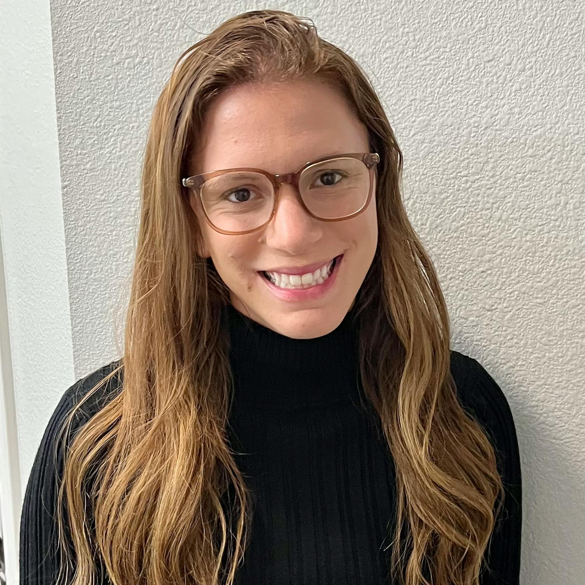Smiling woman with long light brown hair and glasses, wearing a black long-sleeve top, standing against a light-colored textured wall.