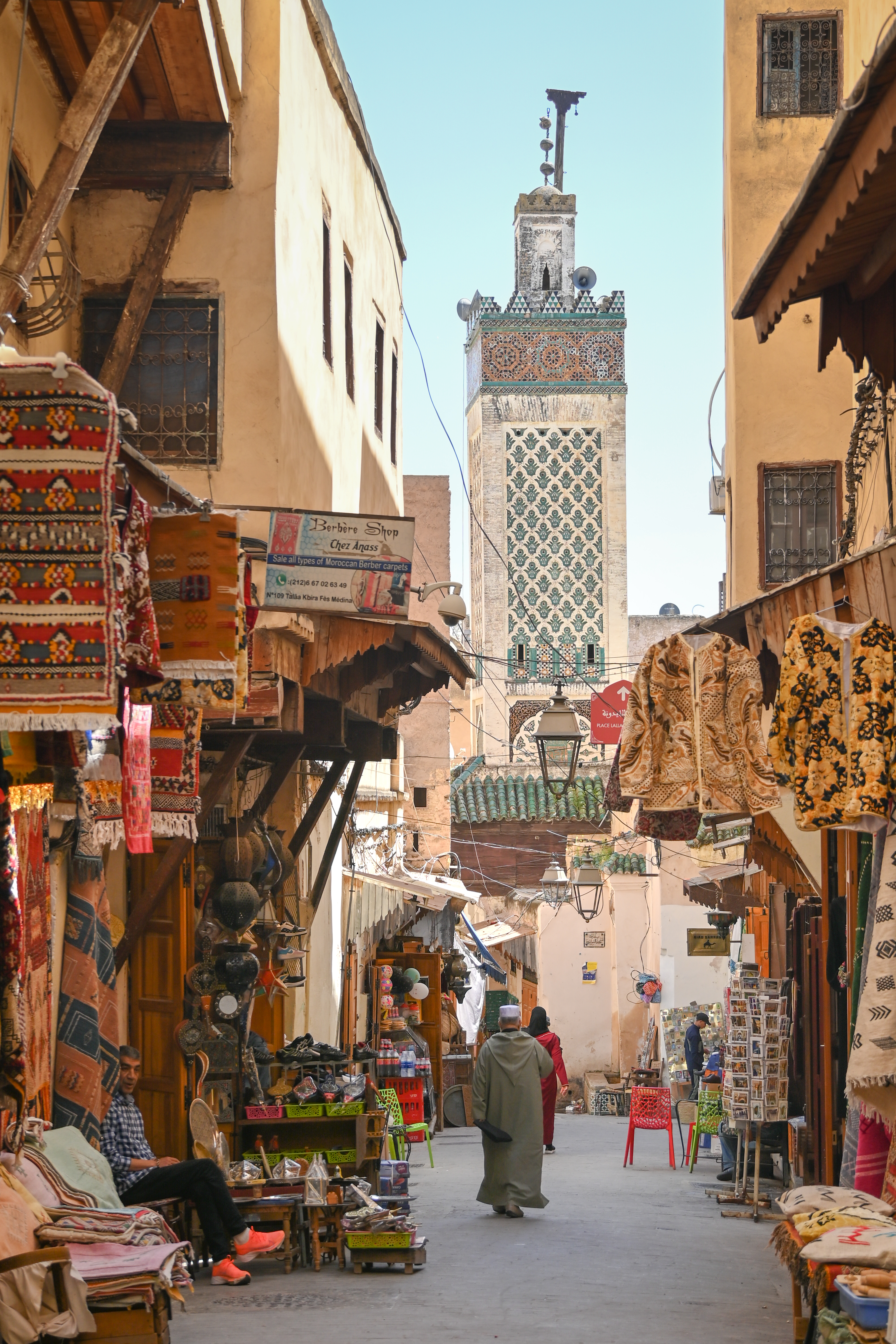 Narrow street lined with shops and pedestrians, with a tall tower in the distance.