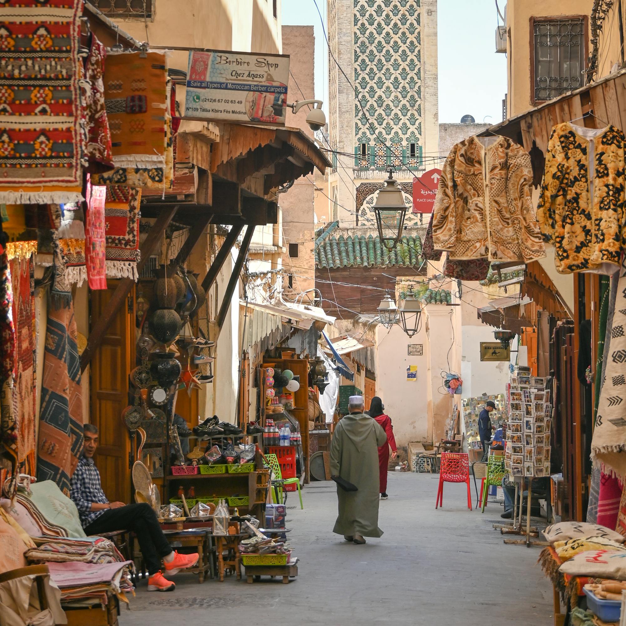 Narrow street lined with shops and pedestrians, with a tall tower in the distance.